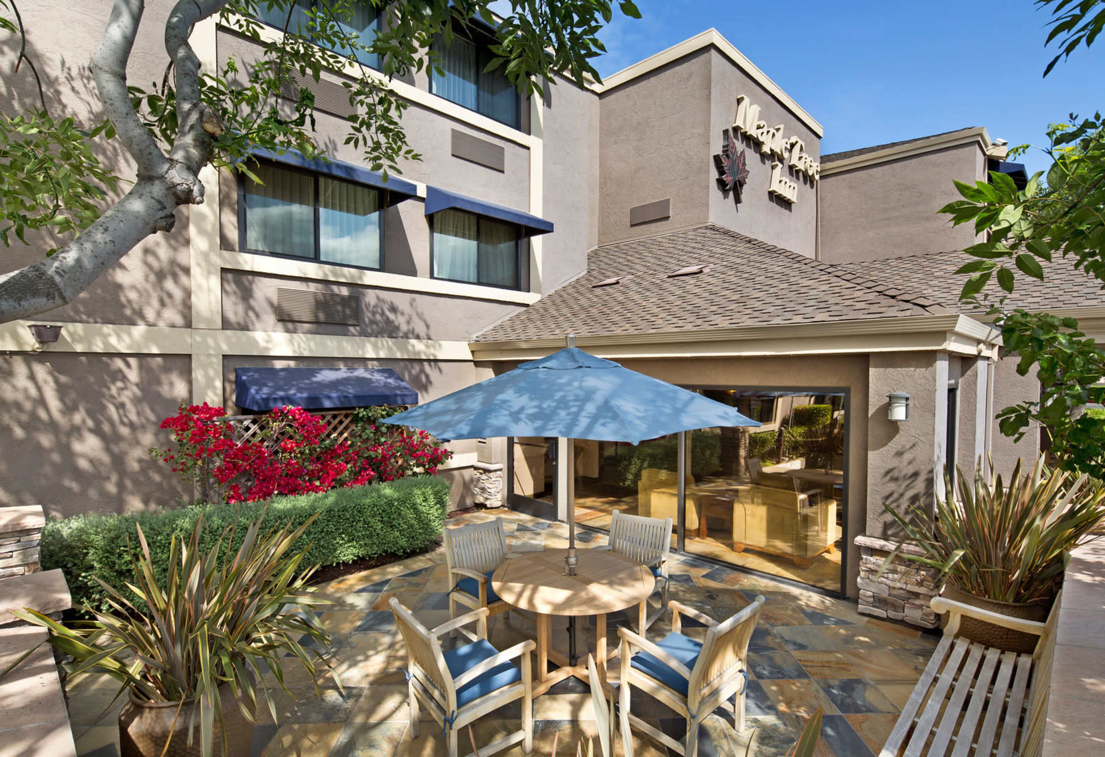 The image shows a patio area with a table and chairs under a blue umbrella, surrounded by greenery and flowers, next to a hotel building.