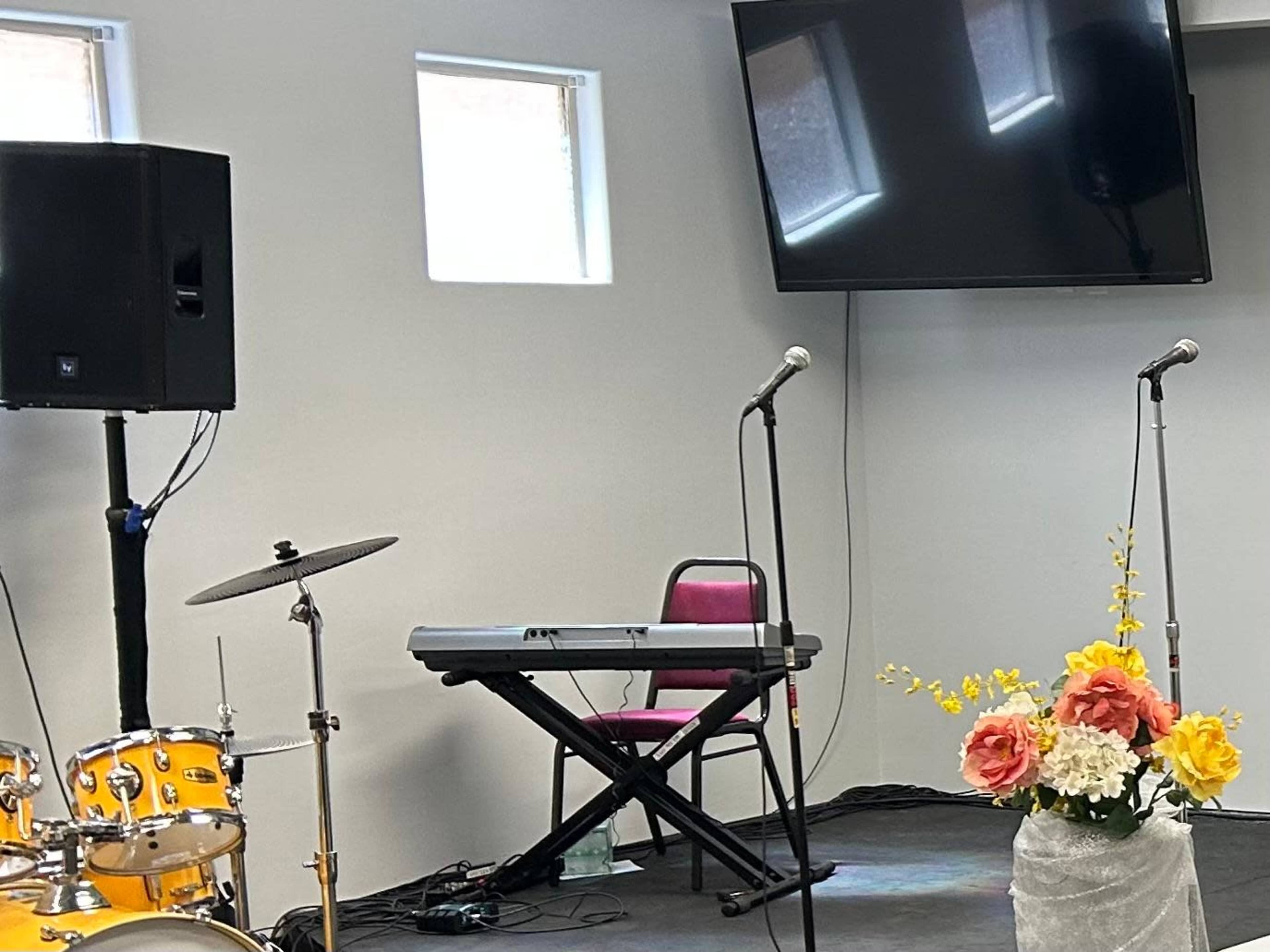 The scene features a musical setup with a drum kit, keyboard, two microphones, a TV screen, and a floral arrangement on a stage.