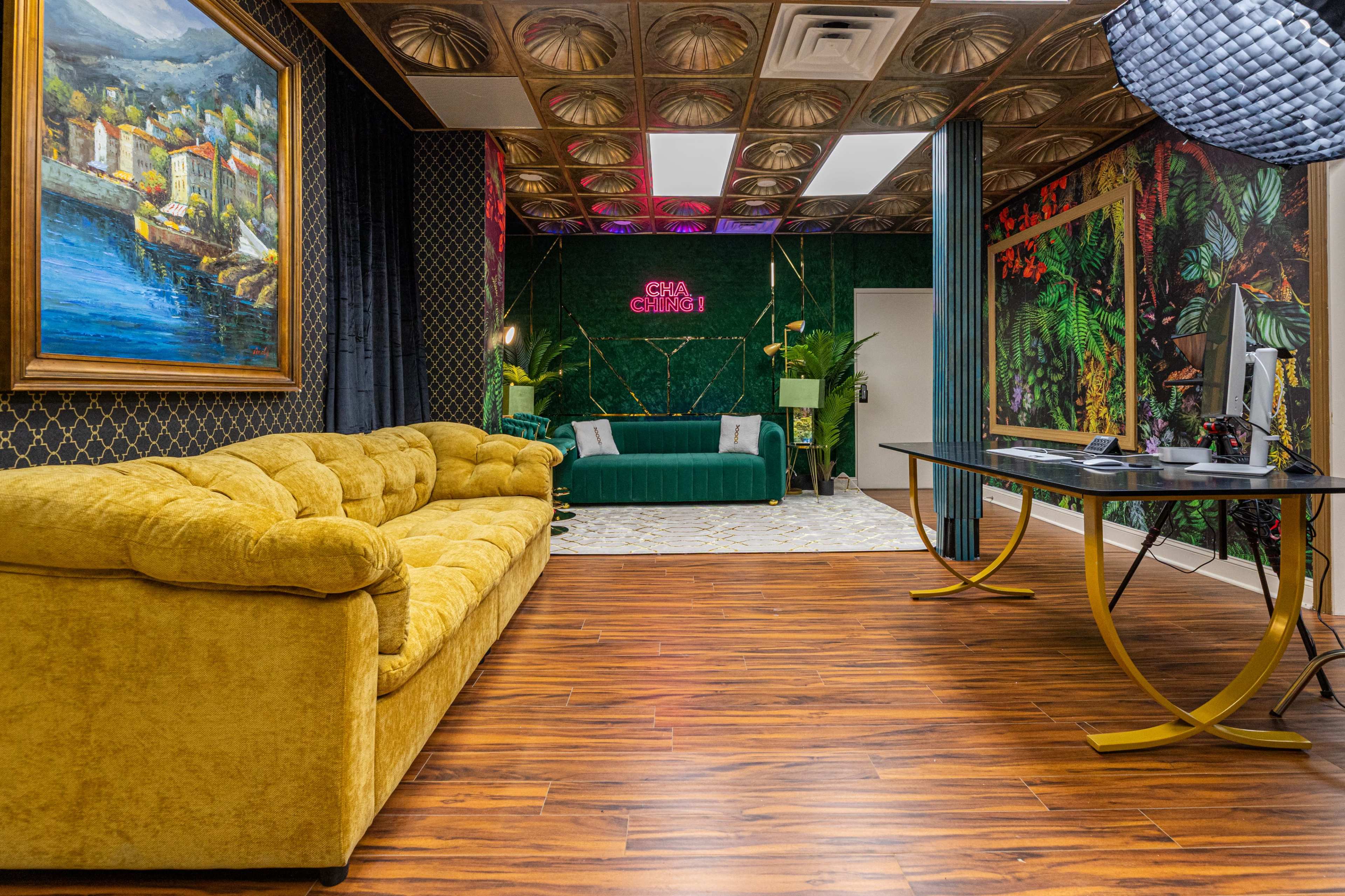 The image shows a stylish interior with a yellow velvet couch, a green couch in the background, plants, and decorative walls featuring tropical patterns.