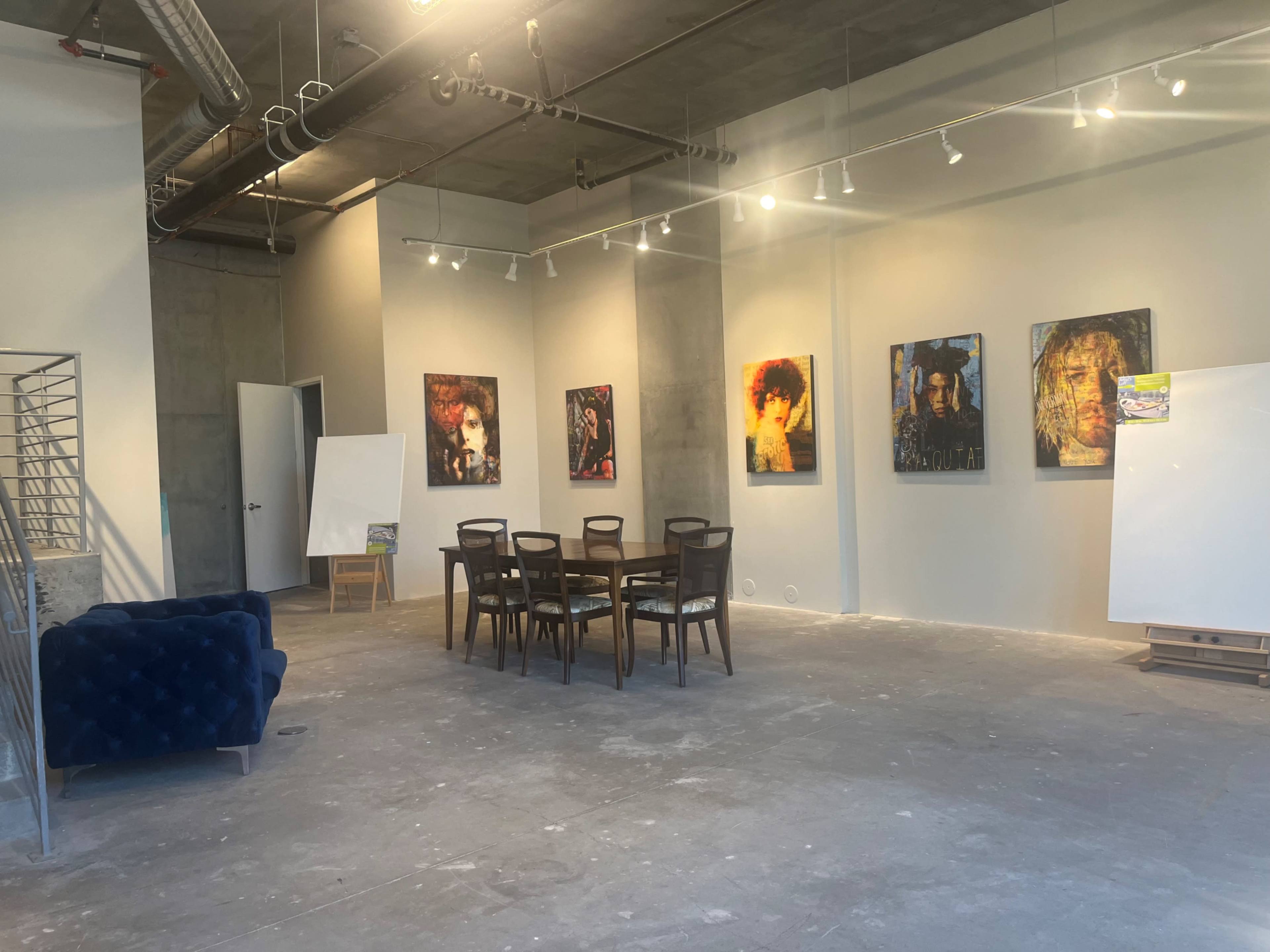 A spacious, unfinished gallery with a dining table and chairs, a blue couch, and several artworks hanging on the walls.