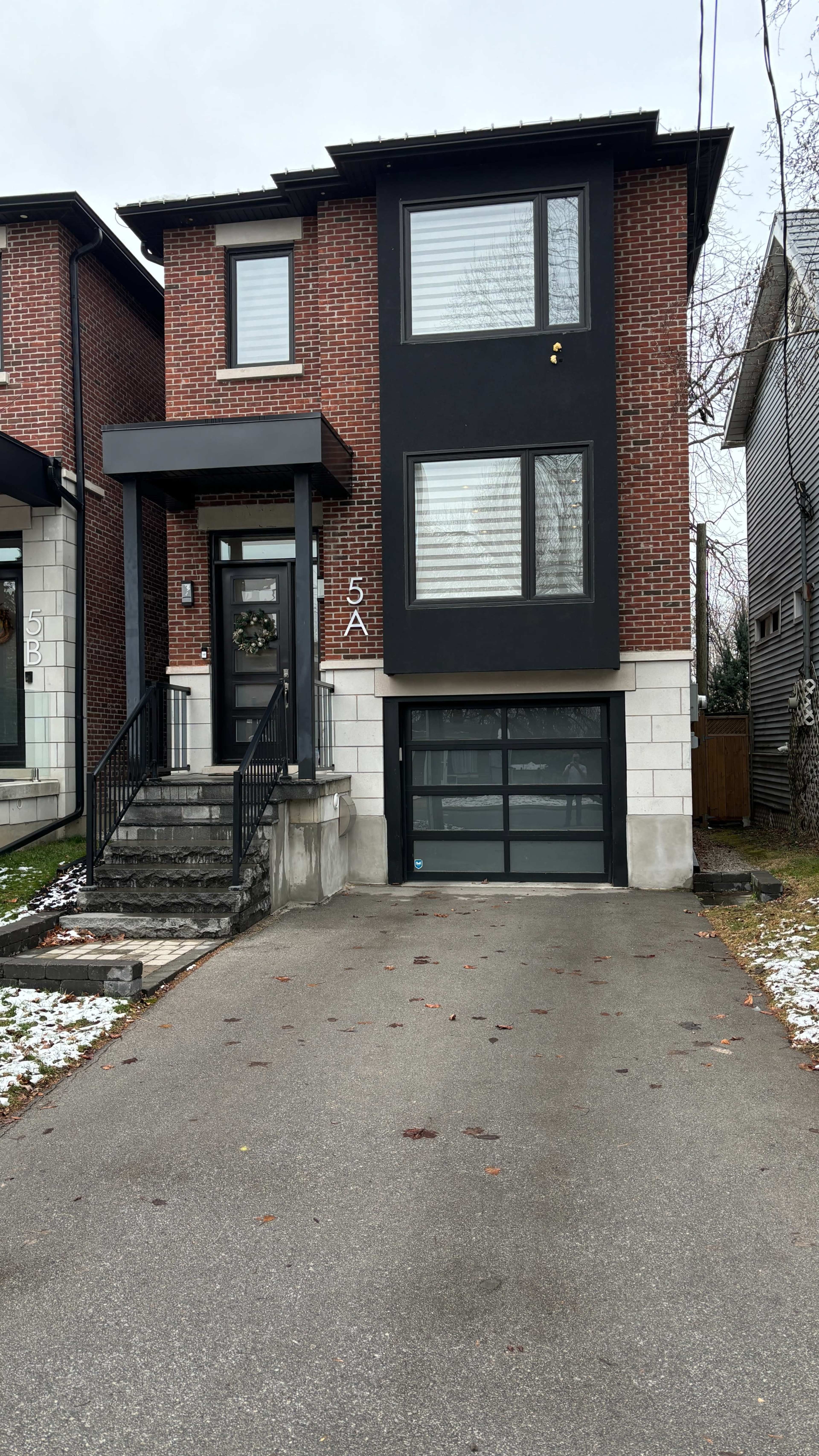A modern two-story brick house with a black exterior and a garage door, set on a residential street with a partially snow-covered driveway.