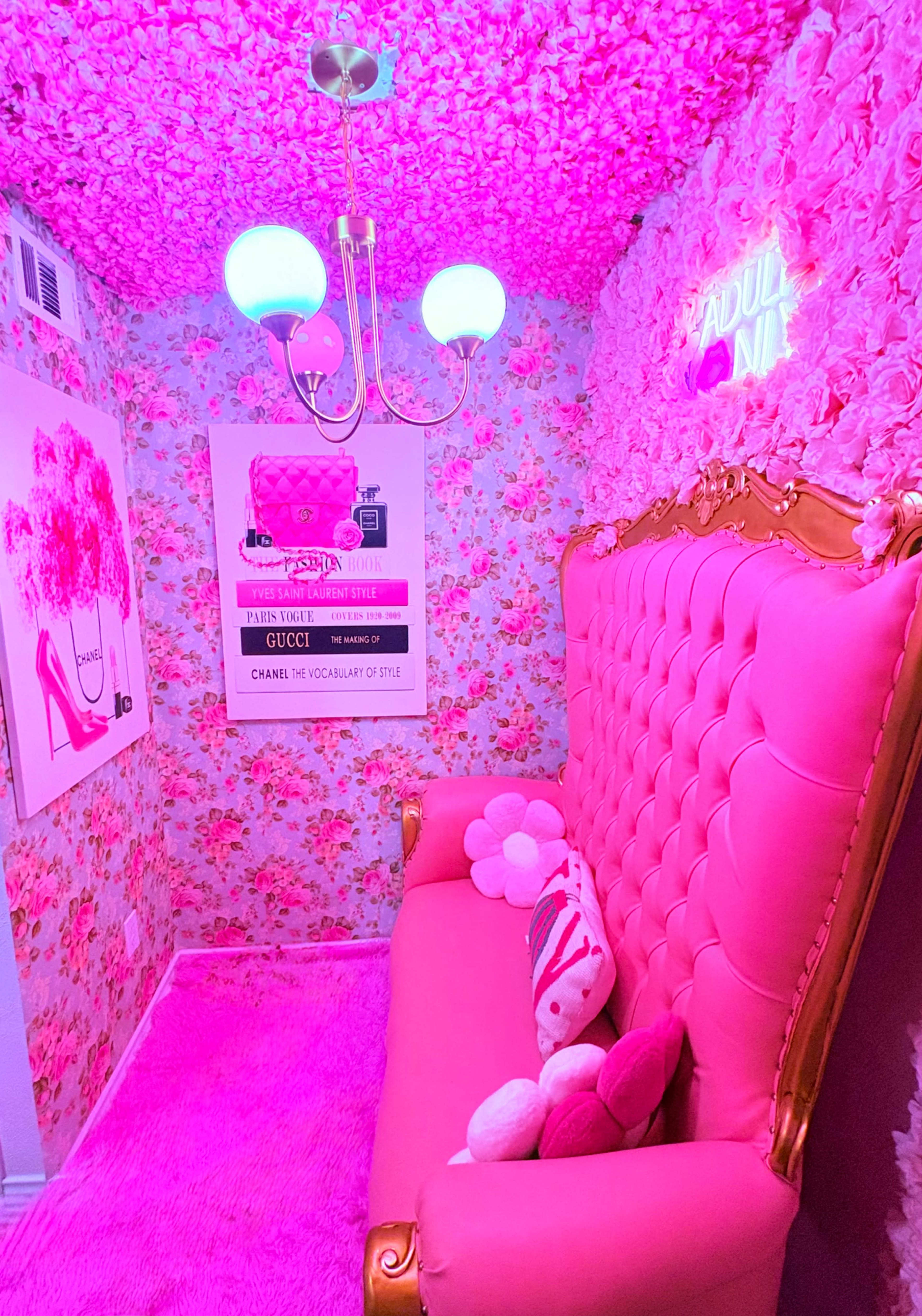 The image shows a small, brightly decorated room featuring a pink velvet couch, floral wallpaper, and a chandelier with glowing bulbs.