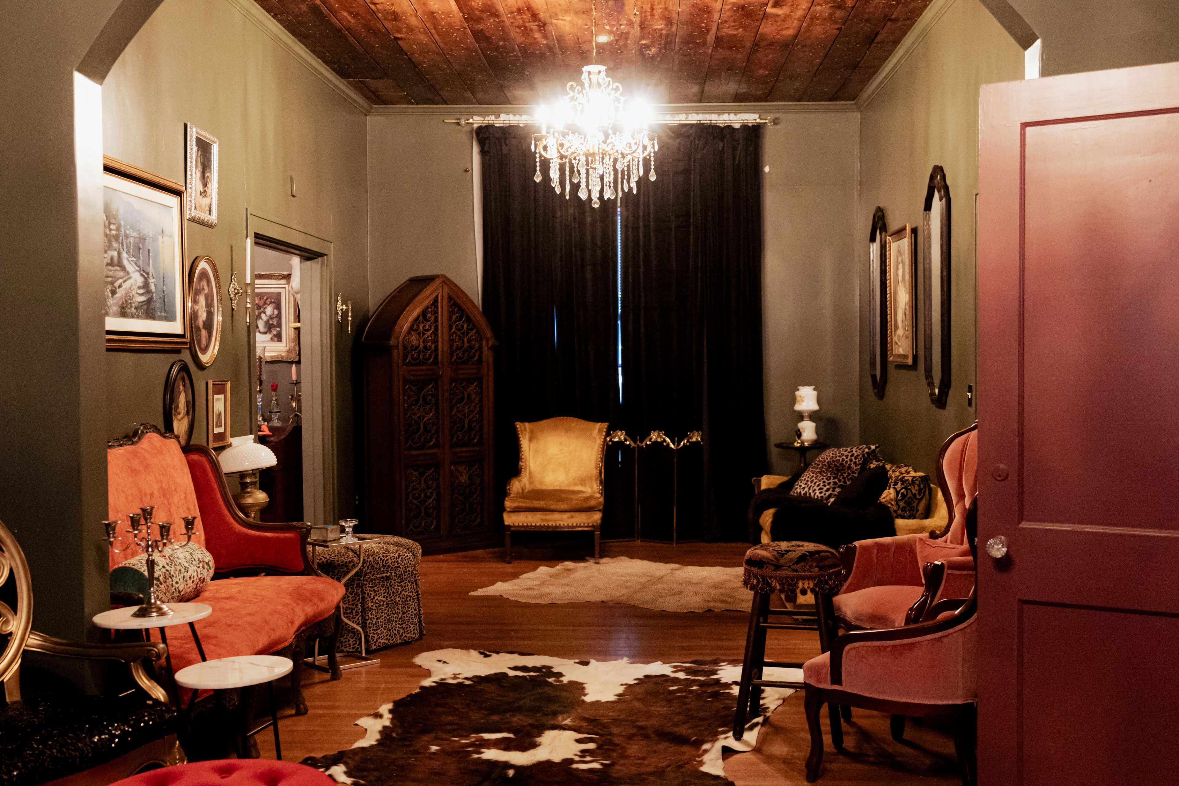 The room features various vintage-style chairs and furniture, illuminated by a chandelier, with a cowhide rug on the wooden floor and dark curtains covering the window.