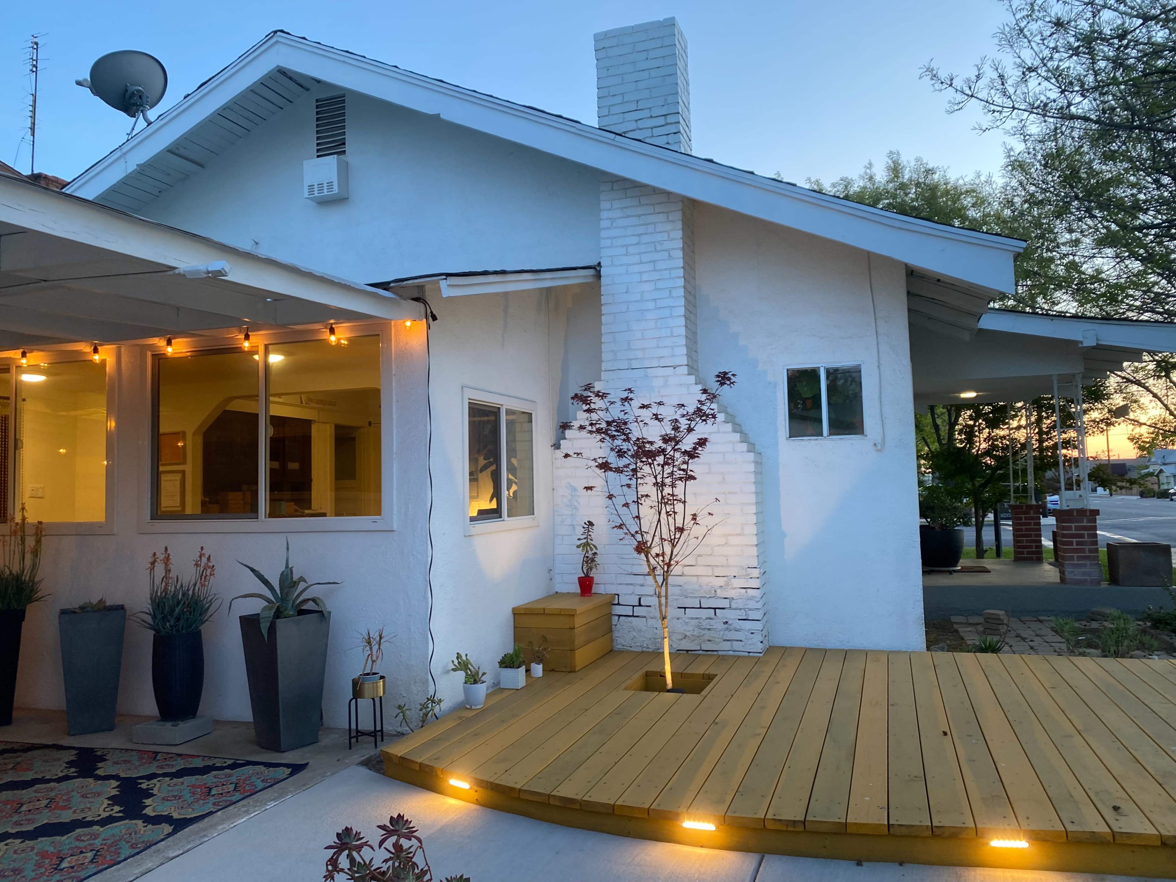 A residential house with a white brick chimney, a wooden deck, and outdoor lighting, situated at the corner of a street.
