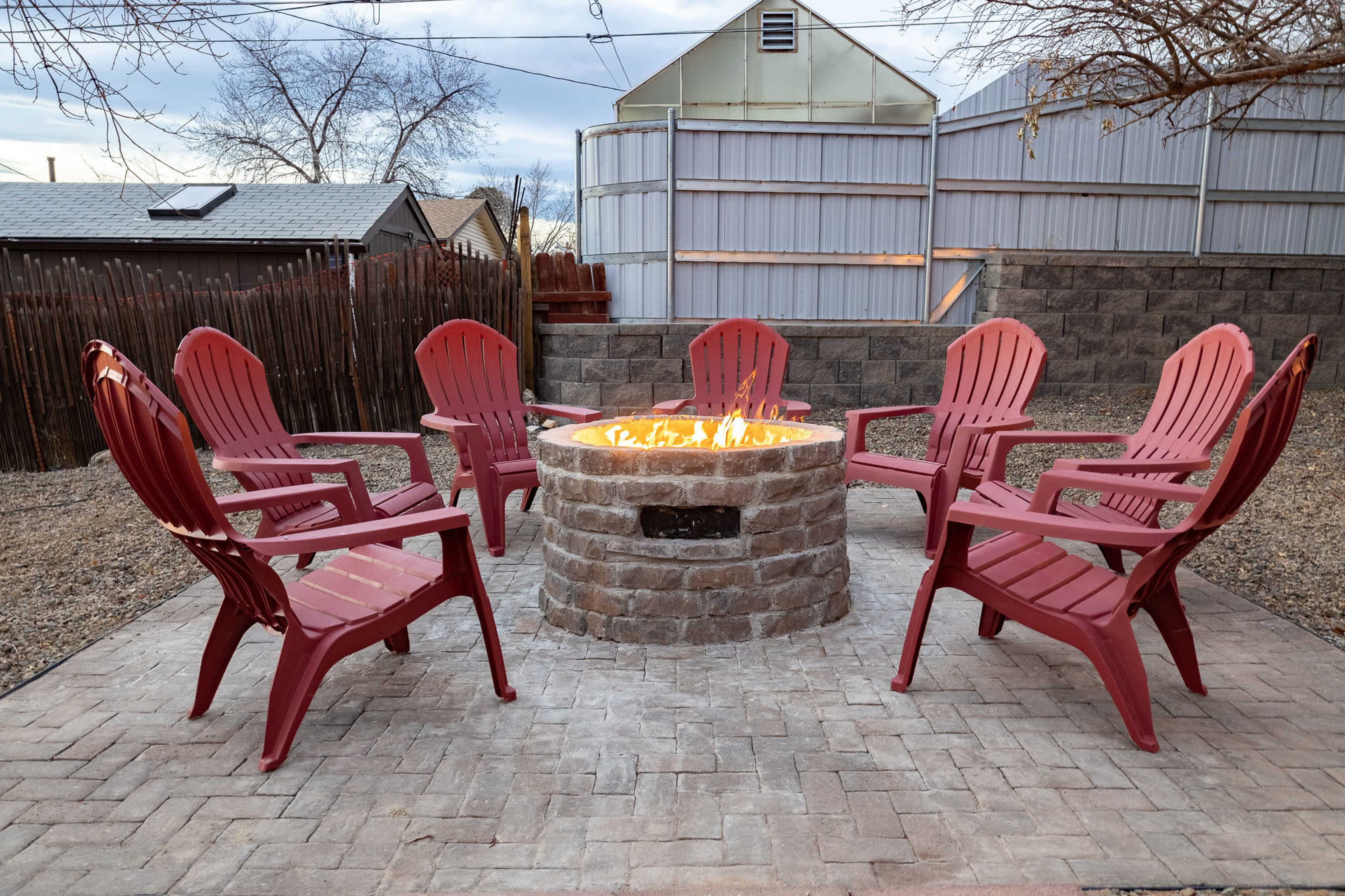 A circular stone fire pit is surrounded by six red Adirondack chairs on a paved patio in a backyard setting.