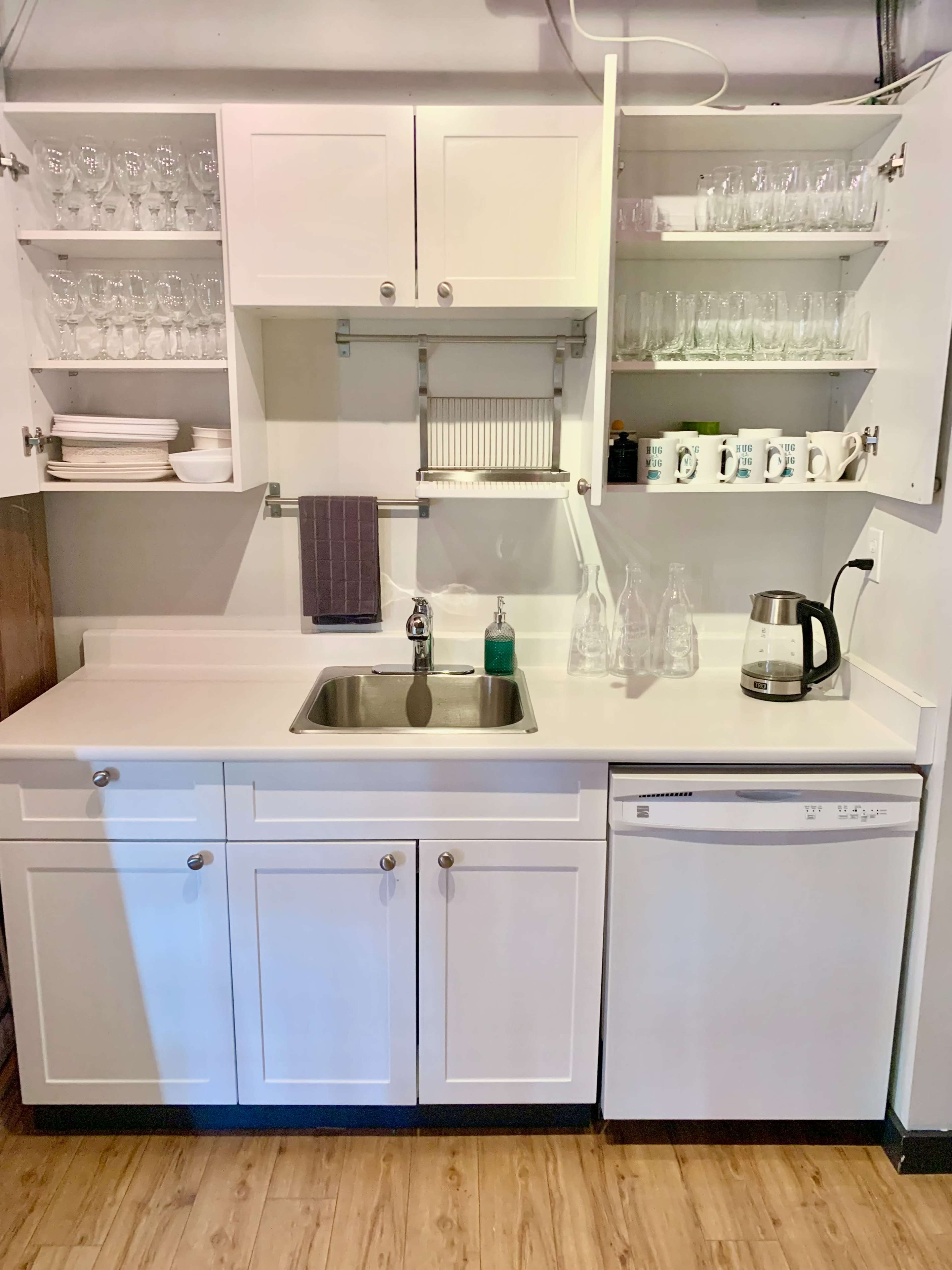 A compact kitchen area with a sink, countertop, dishwasher, and upper shelves filled with glassware and dinnerware.