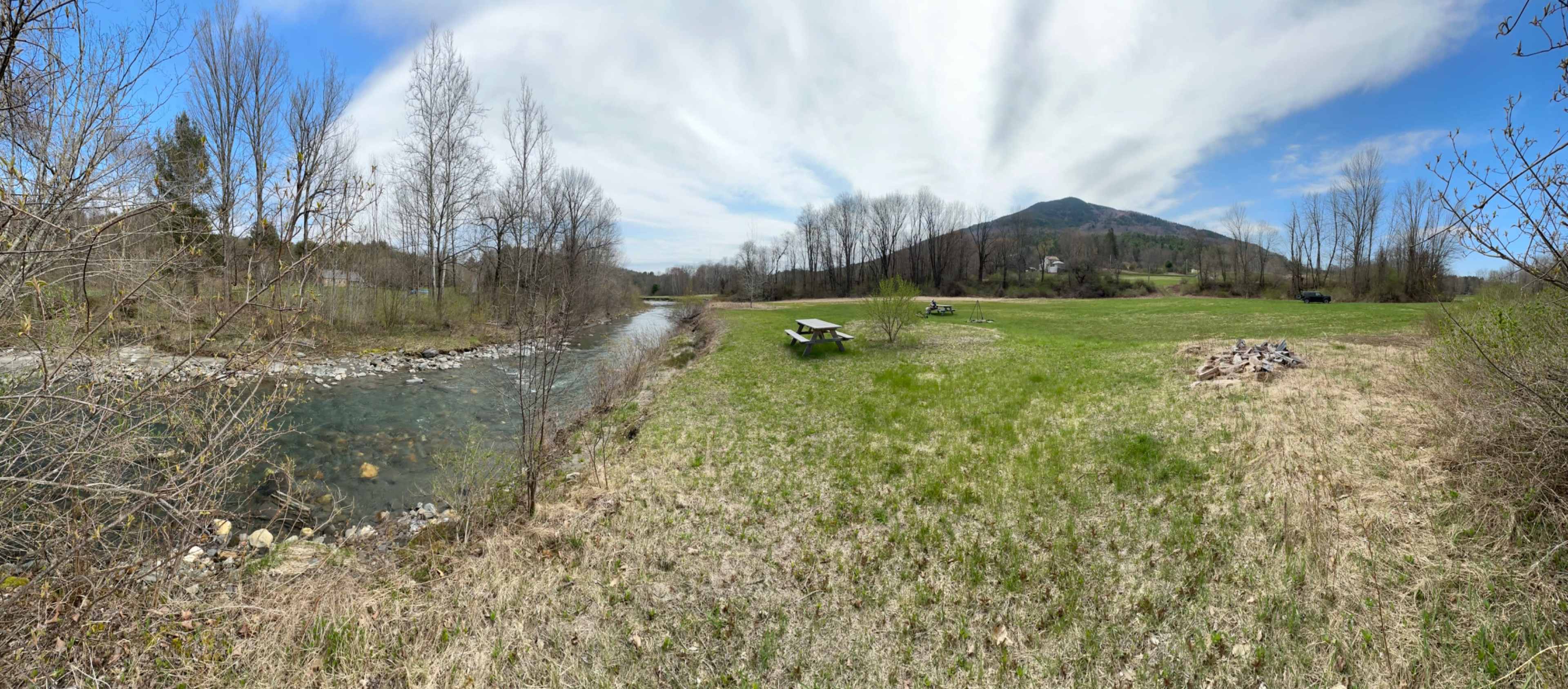 A river flows alongside a grassy area with picnic tables, bordered by trees and a mountain in the background under a cloudy sky.