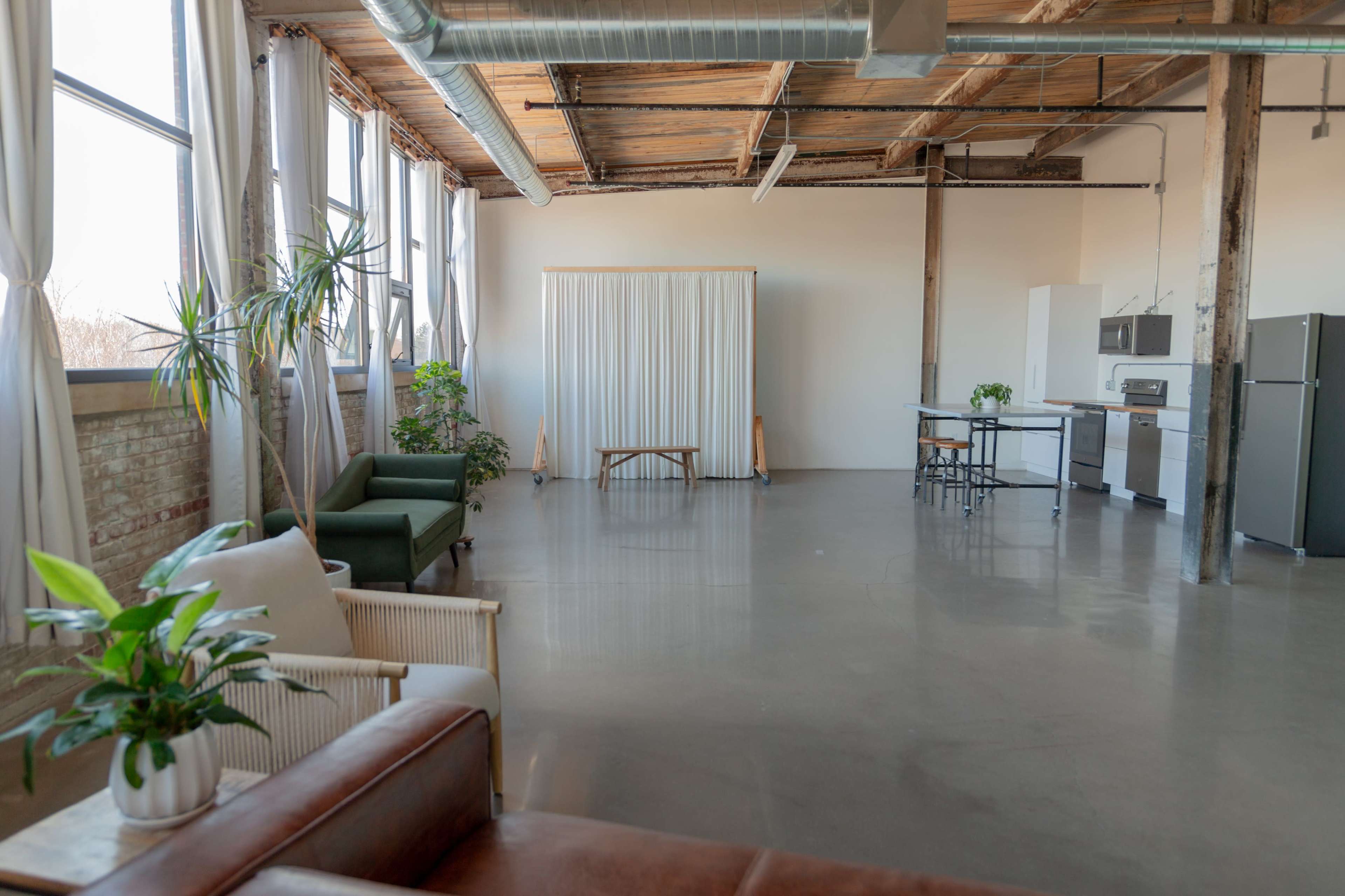 The image depicts a spacious, industrial-style loft with large windows, featuring a minimalist seating area and a simple kitchen setup.