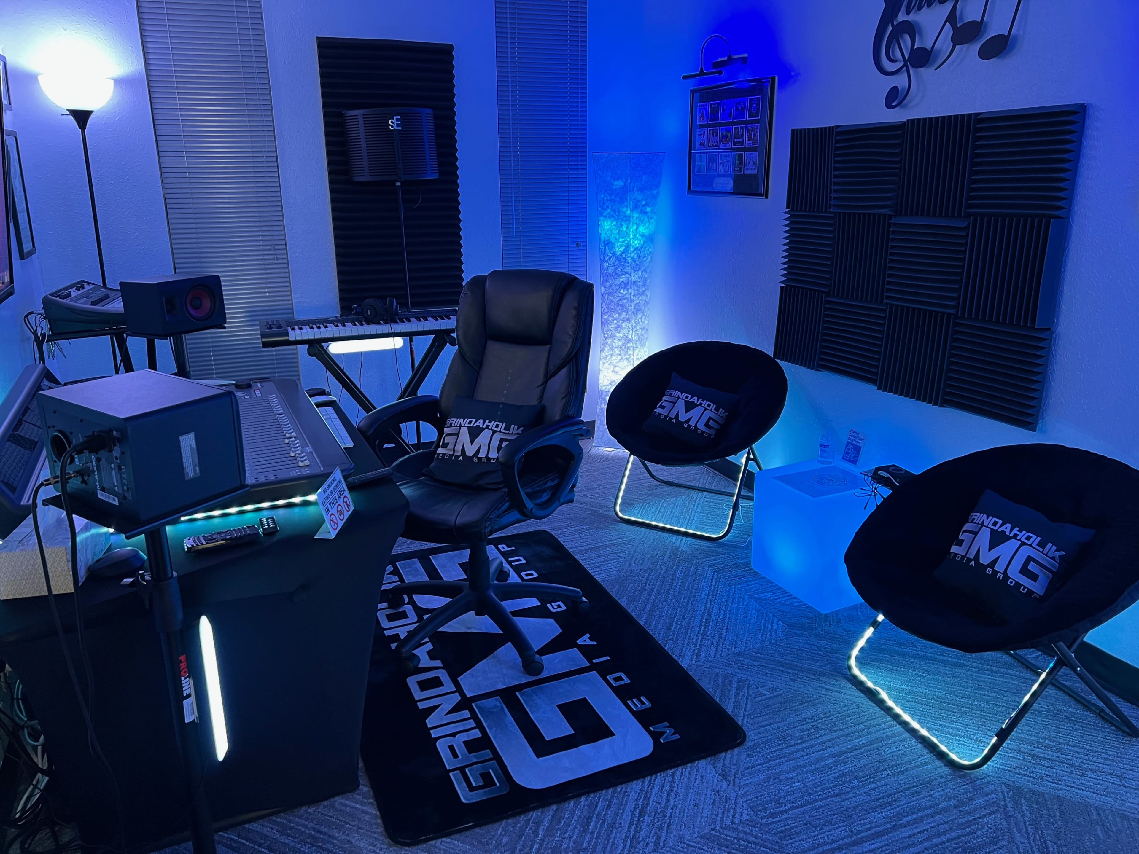 A music studio with a mixing console, keyboard, and two illuminated lounge chairs under blue lighting, along with soundproofing panels on the walls.