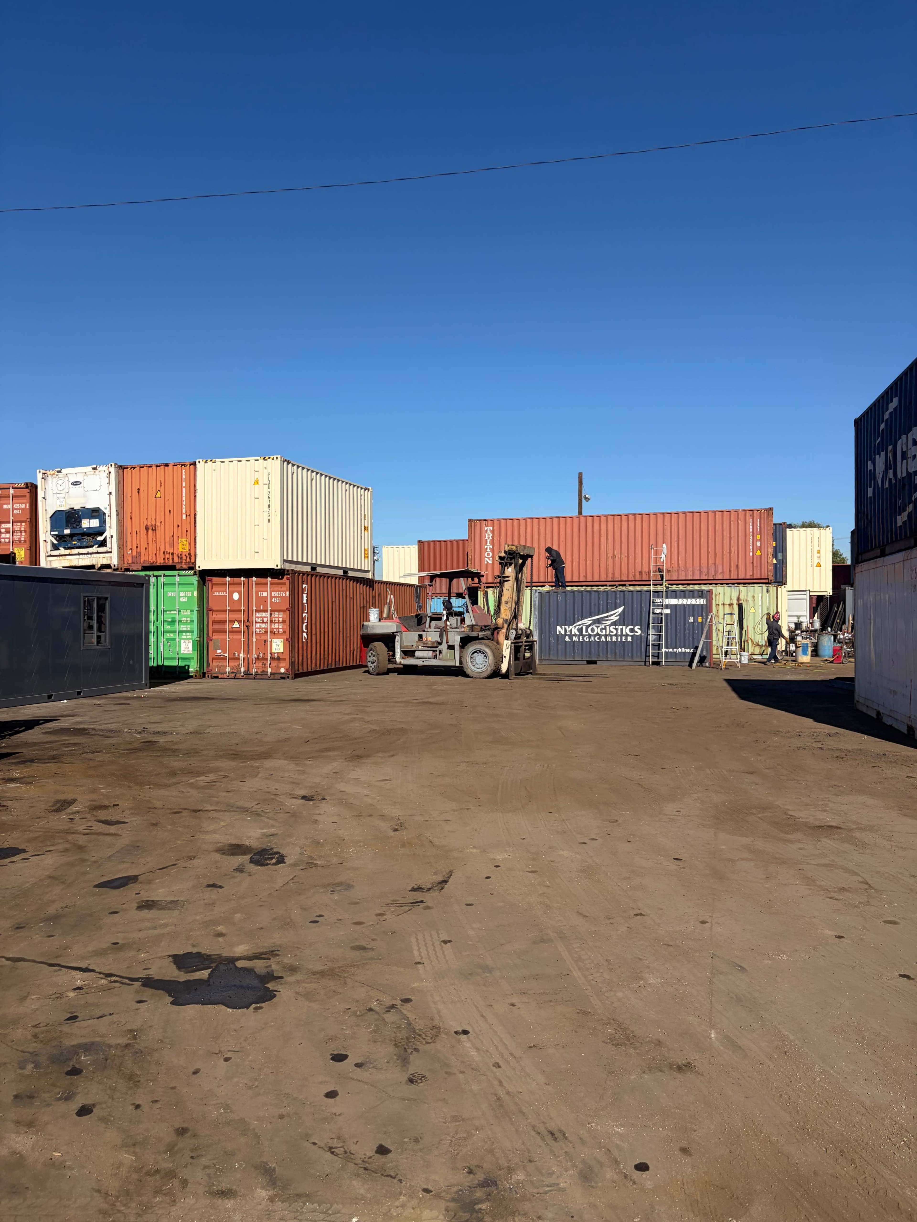 The image shows a storage area filled with stacked shipping containers, with a forklift in the center navigating through the space under a clear blue sky.