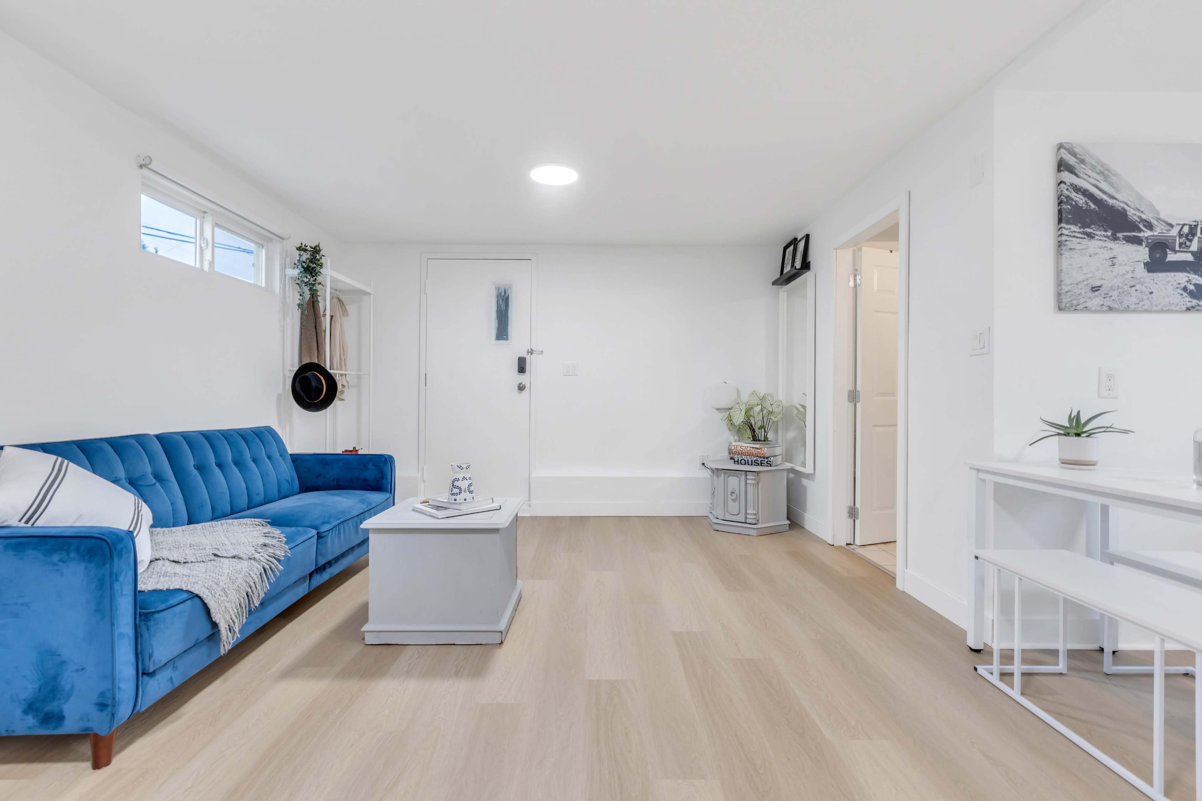 A minimalist living room featuring a blue couch, a light-colored coffee table, and a white console table against pale wood flooring.
