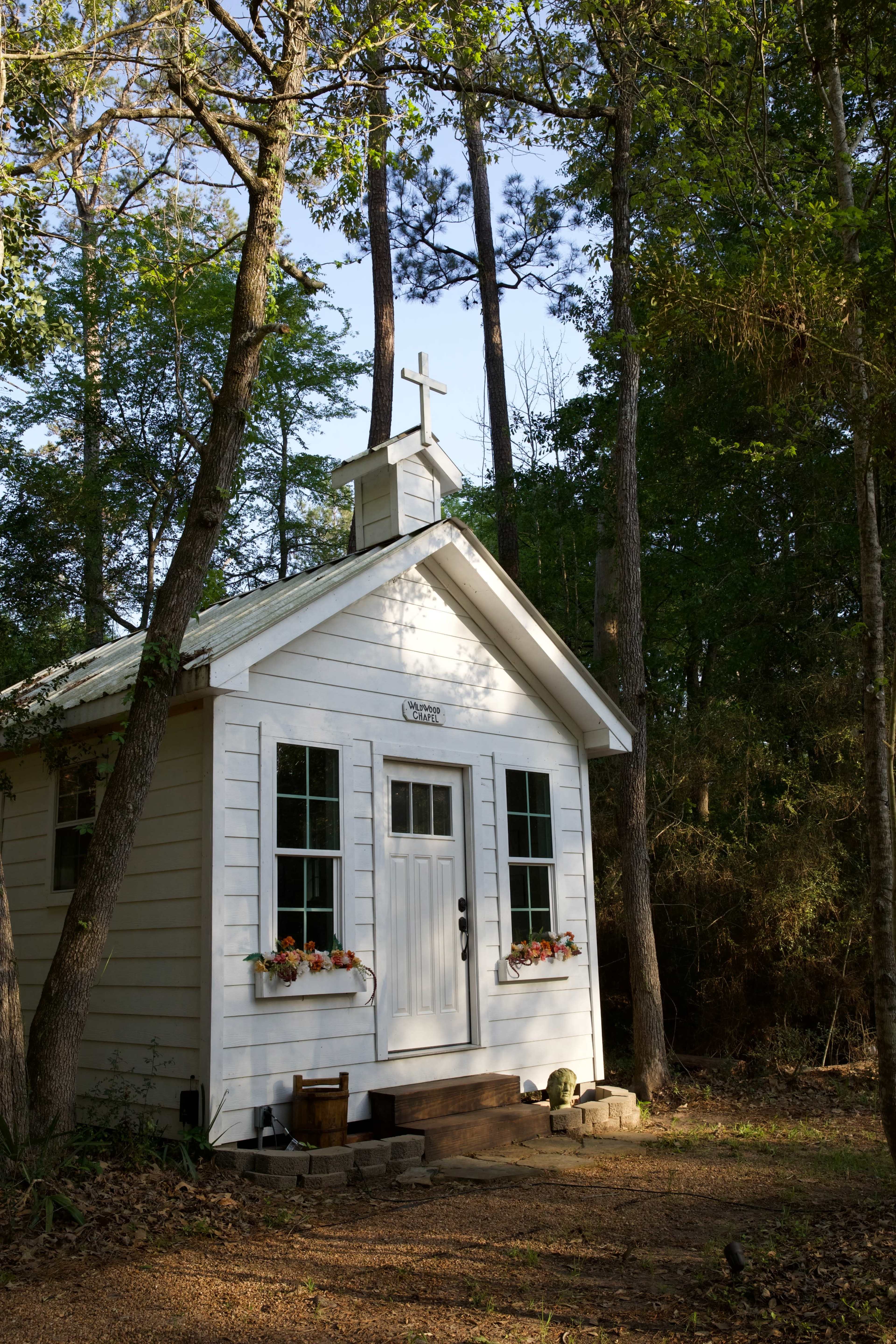 A small white wooden chapel with a cross on top is surrounded by trees in a wooded area.