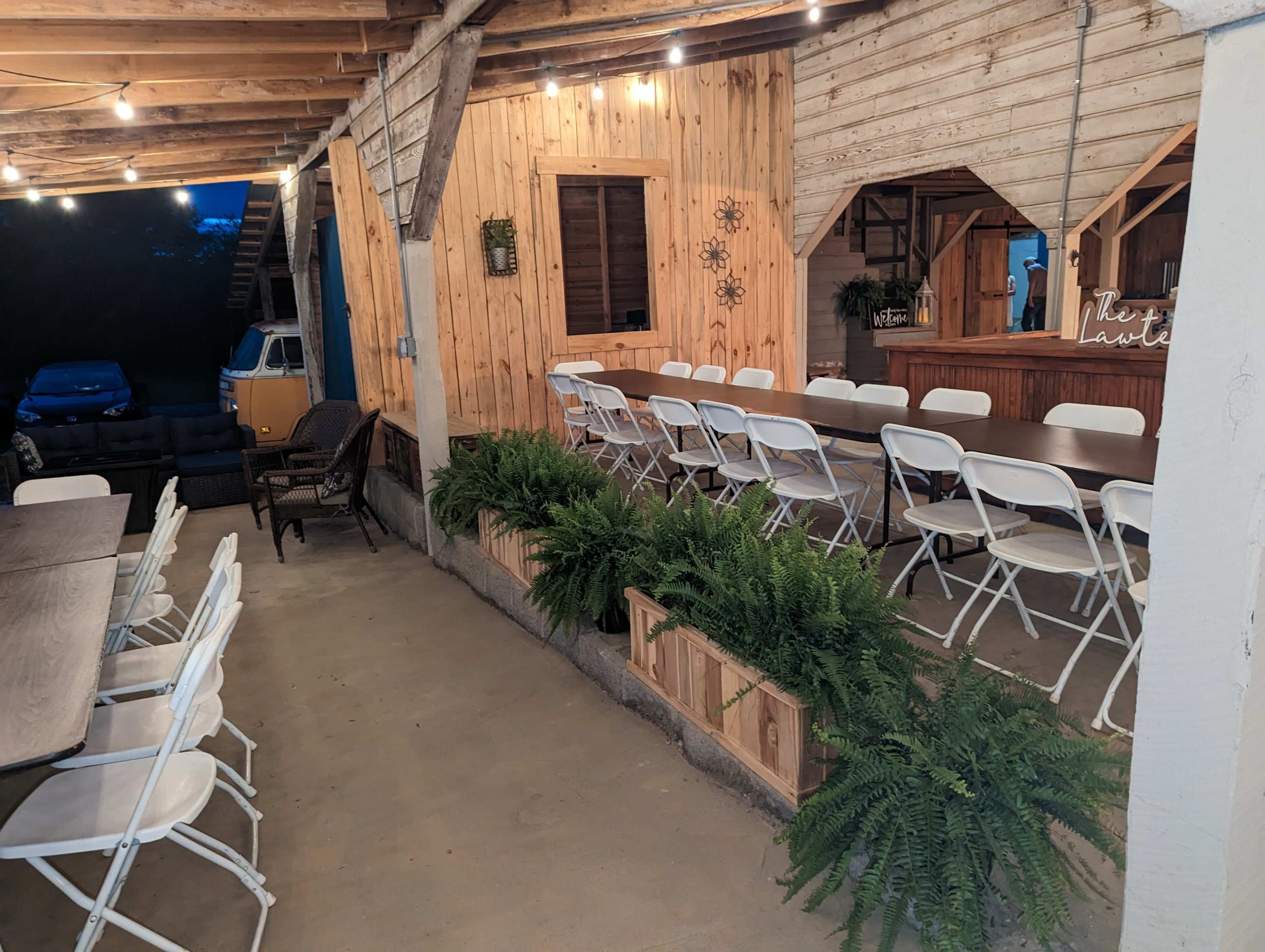 An outdoor covered area with several white folding chairs arranged around tables, surrounded by wooden walls and planters filled with ferns.