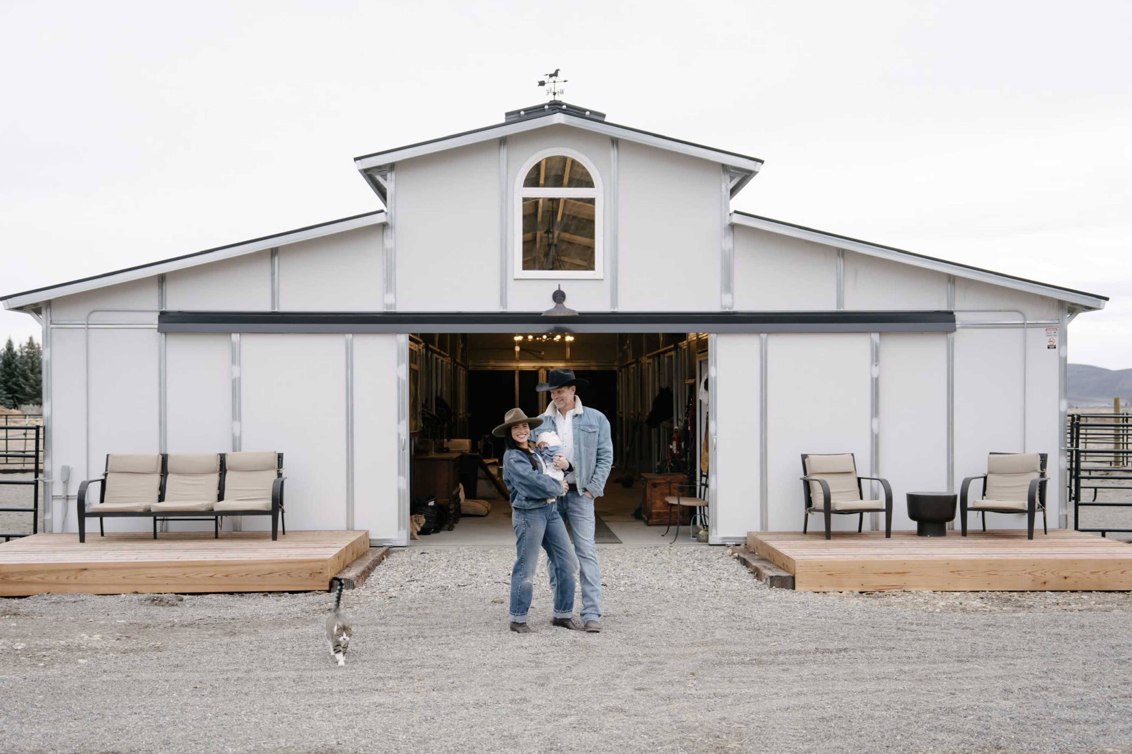 A woman and a child stand in front of a spacious barn with an open entrance, flanked by outdoor seating on wooden platforms.