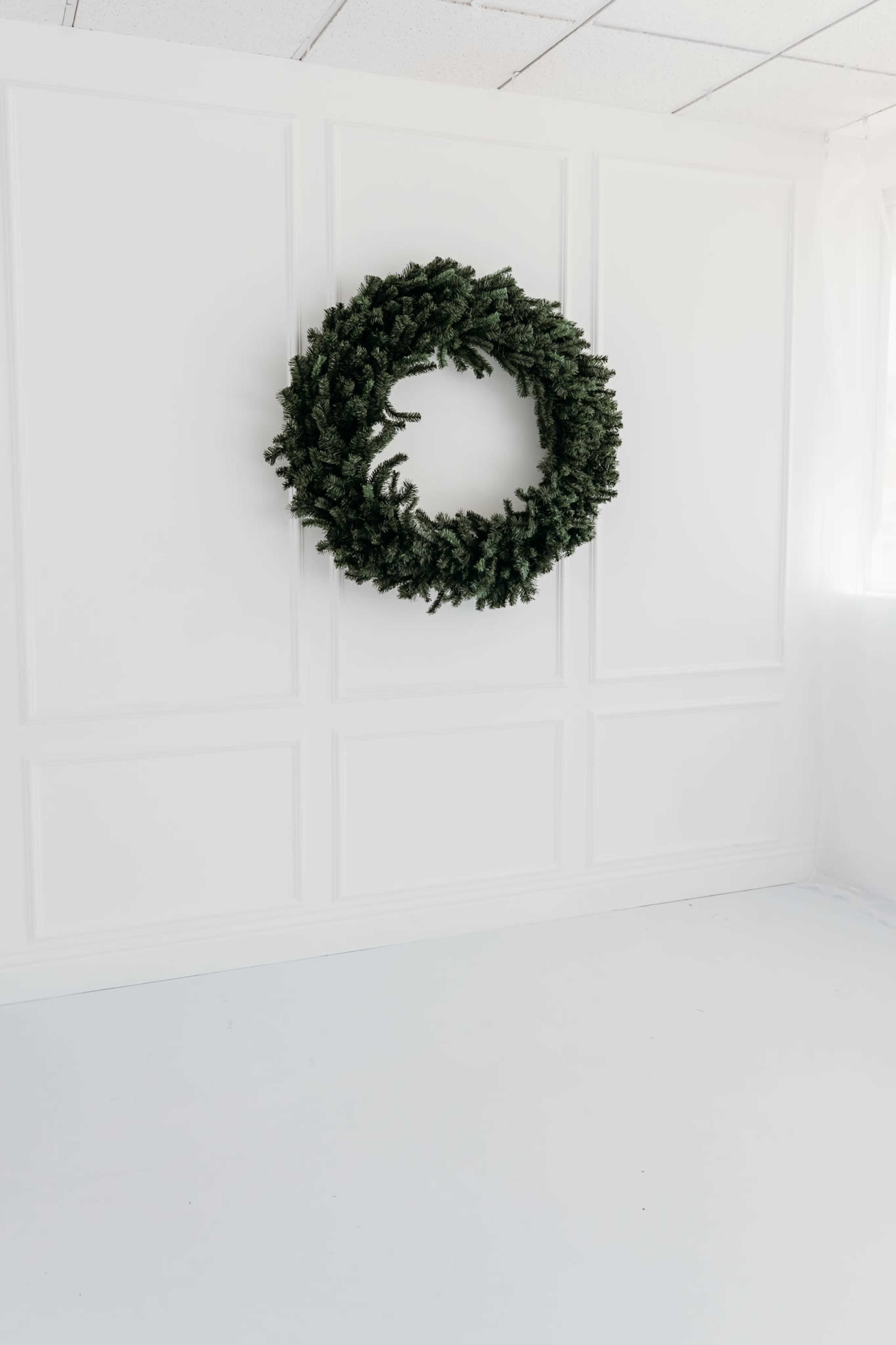 A large, green wreath is mounted on a white paneled wall in a spacious, bright room with a white floor.