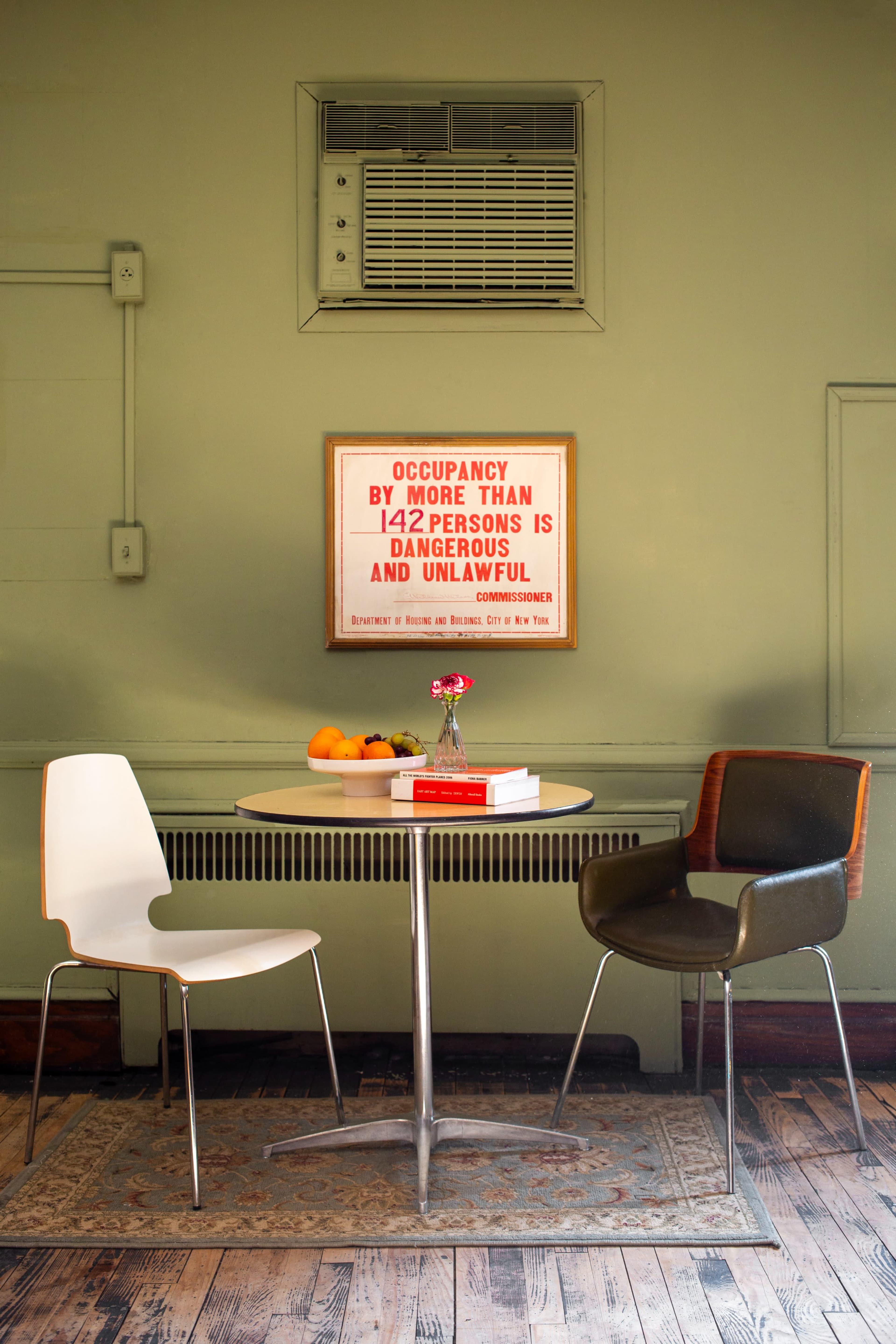 A small dining table with a white chair and a dark green chair sits on a patterned rug in front of a wall-mounted air conditioner and a framed sign about occupancy limits.