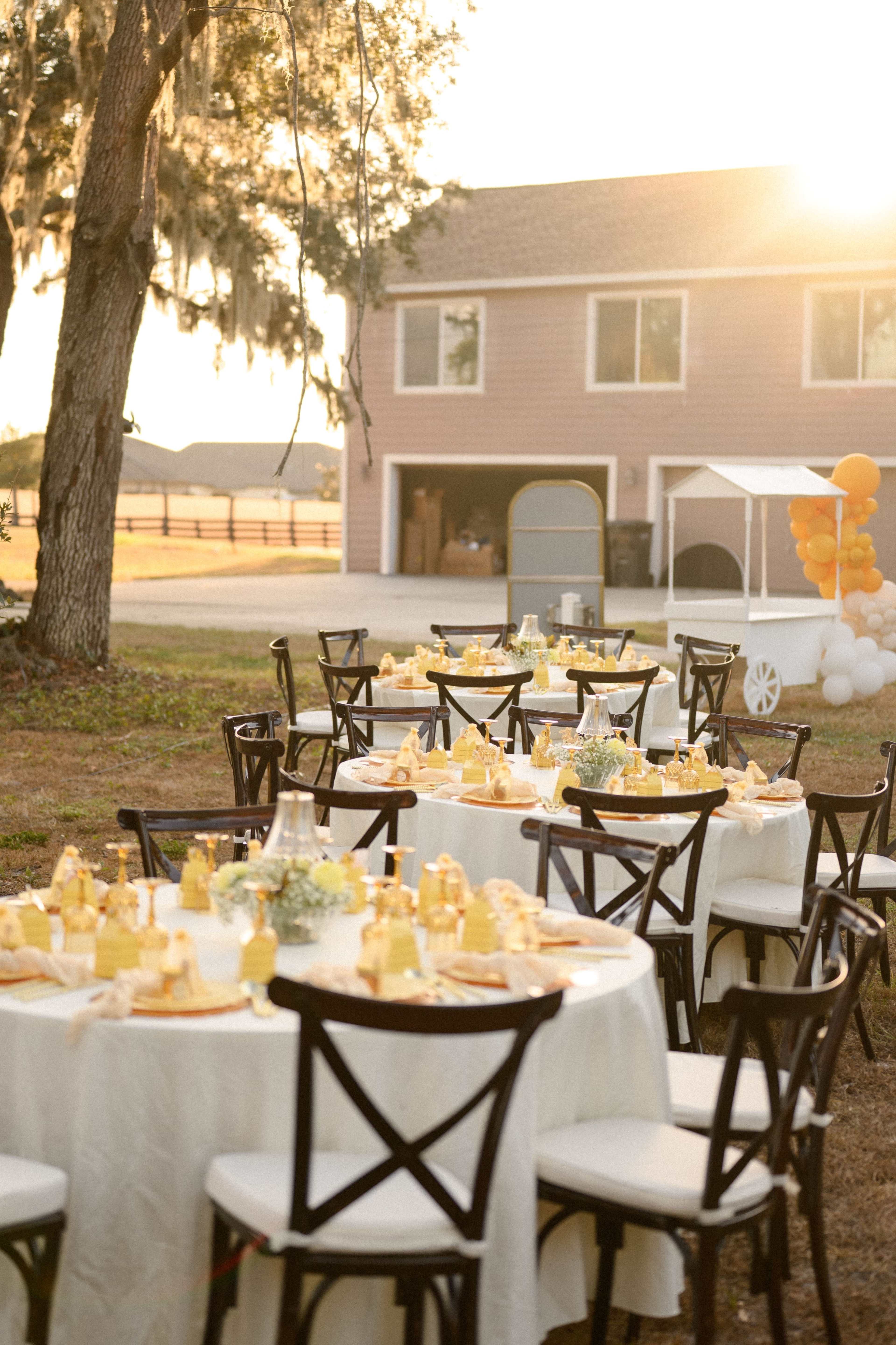 A set of elegantly arranged tables with black chairs is positioned outdoors, featuring yellow-themed decorations, in front of a house and a vintage-style dessert cart.
