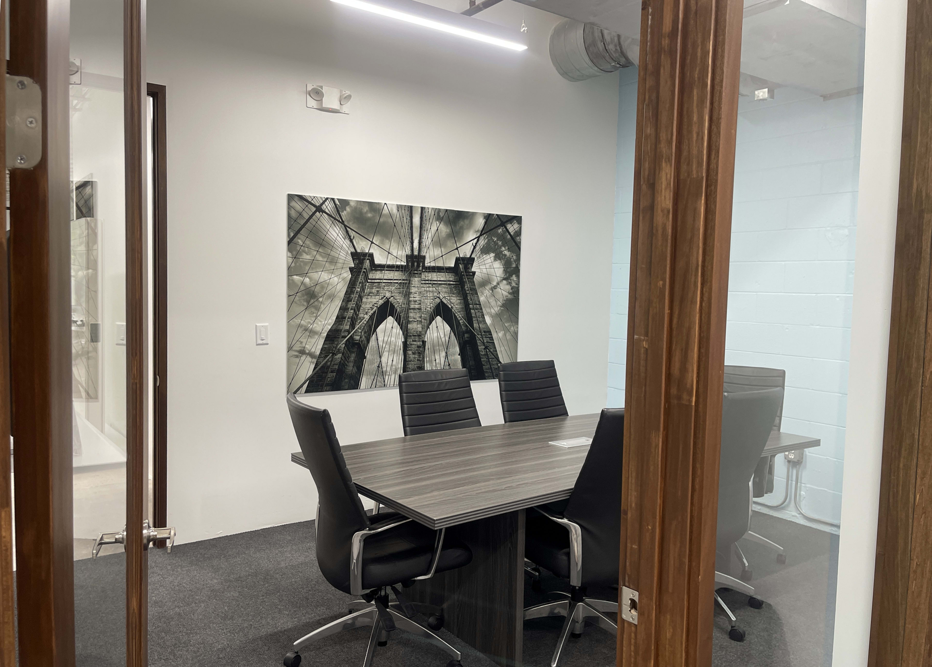 A glass-enclosed conference room features a large photograph of the Brooklyn Bridge on the wall, with a wooden table and black chairs arranged around it.