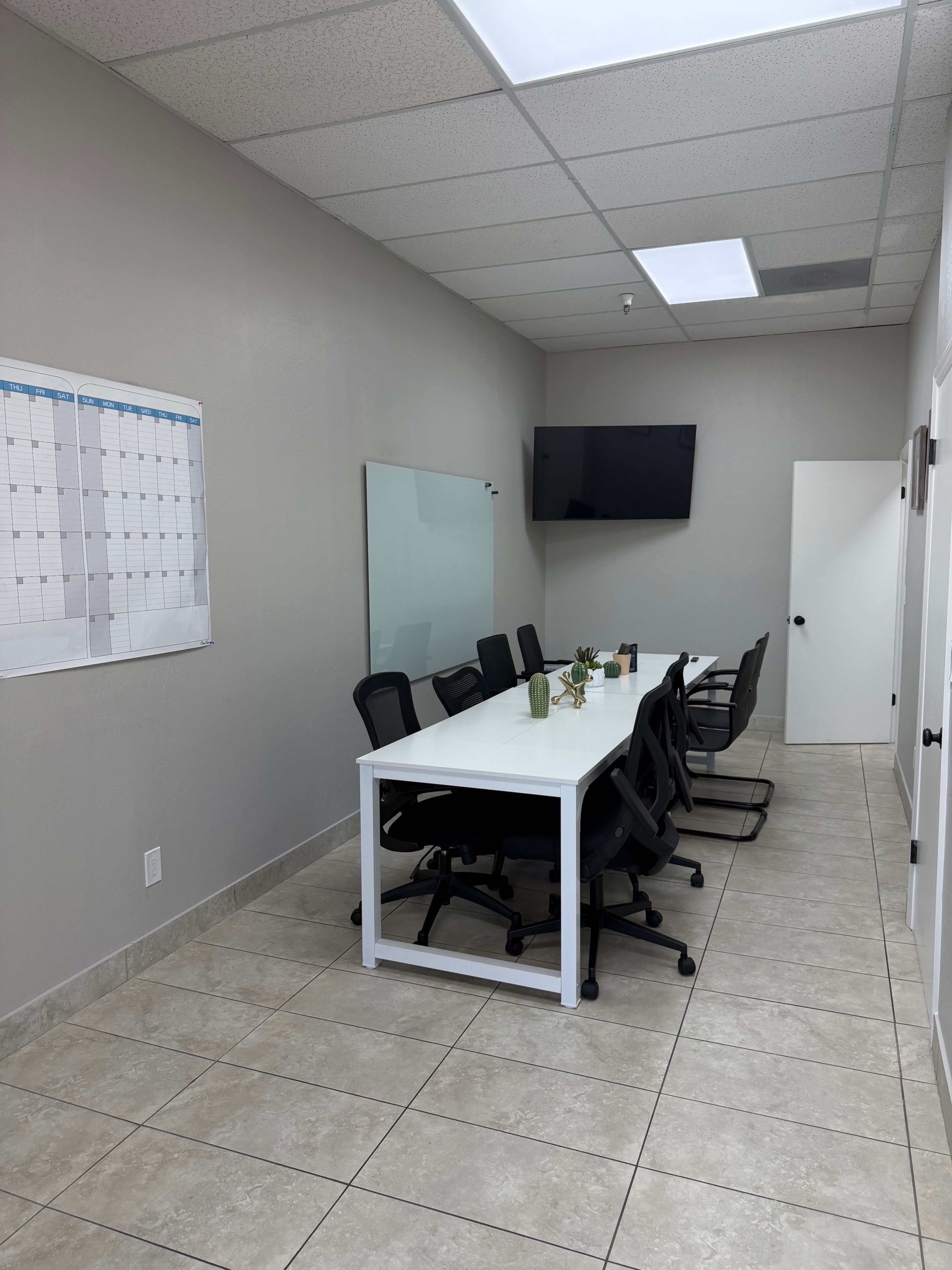 The image shows a modern meeting room featuring a long white table surrounded by black office chairs, with a wall-mounted TV and a calendar on the adjacent wall.