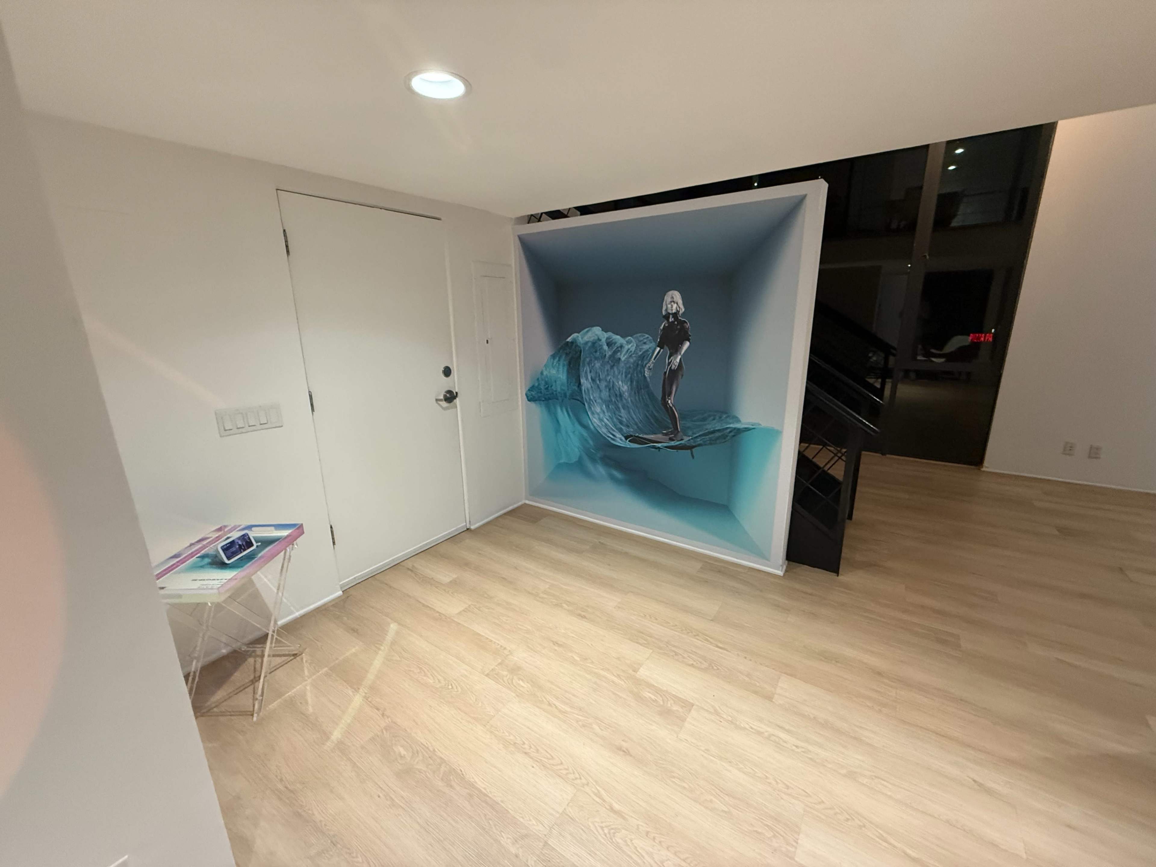 The image shows a modern interior space with light wood flooring, featuring a three-dimensional mural of a surfer riding a wave against a white wall.