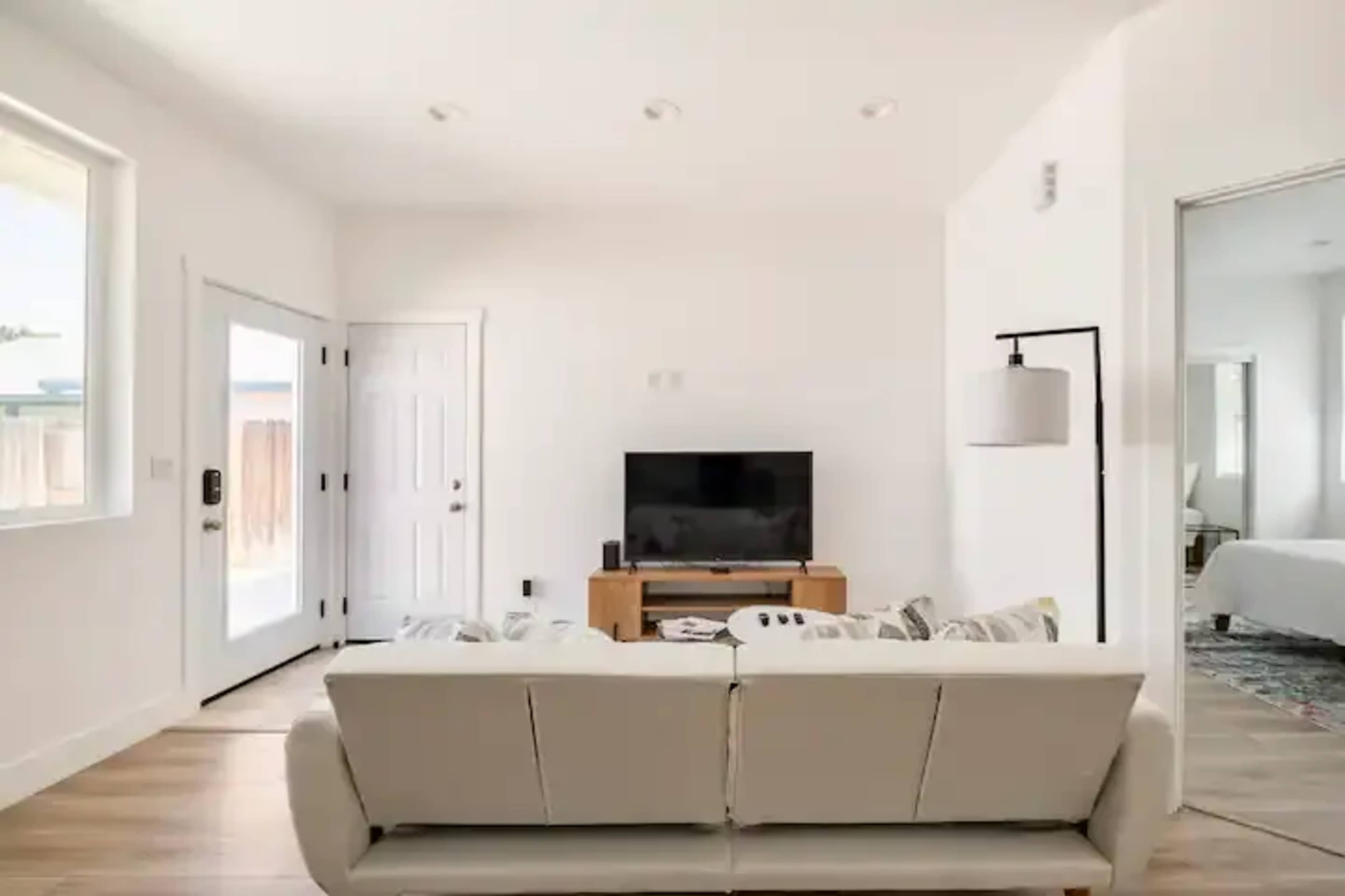 A modern living room features a beige sofa facing a television, with a door leading outside and a light fixture beside it.