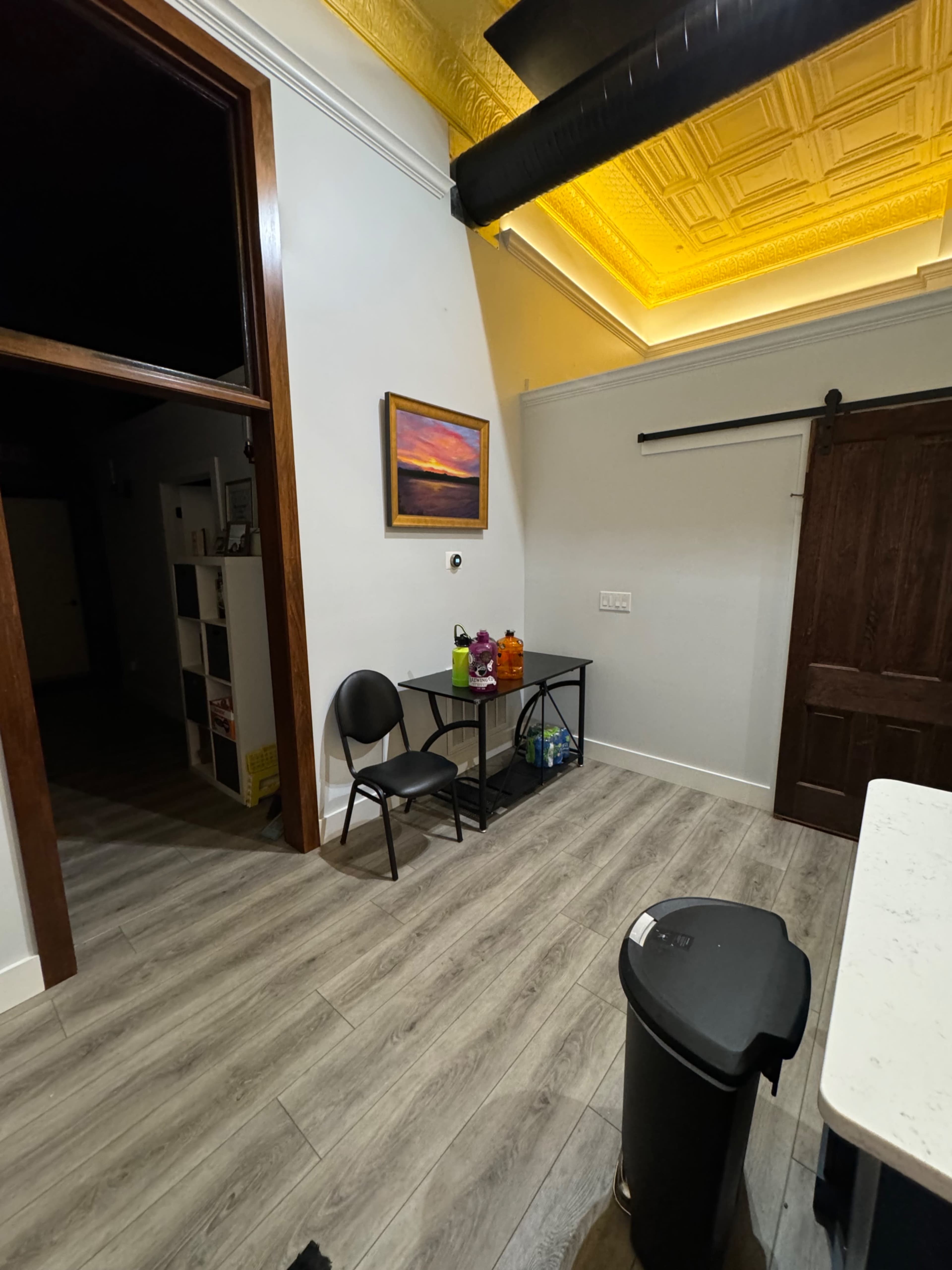 The image shows a room with a small table and two chairs, a waste bin, a door on the right, and a painting on the wall above the table.