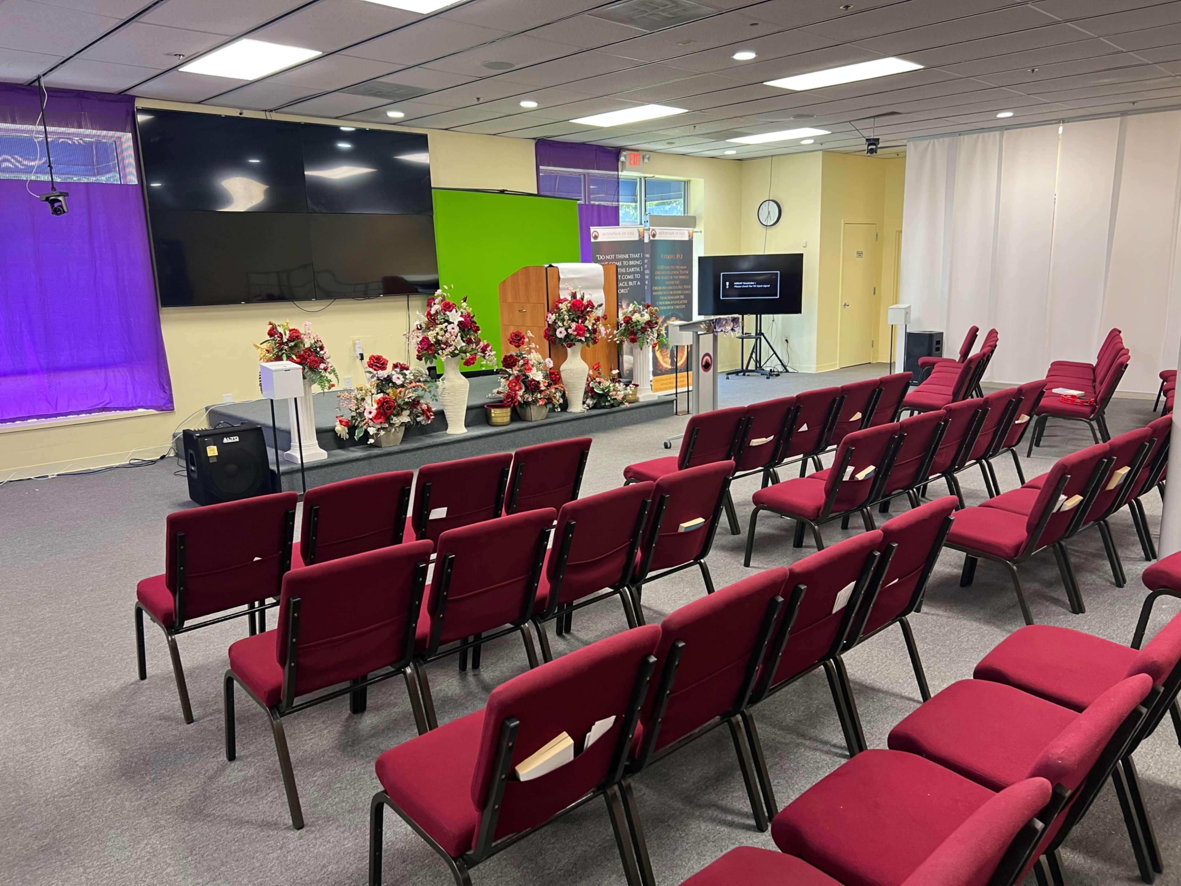 The image shows a seminar room with rows of red chairs facing a stage decorated with flower arrangements and a large screen, with a green backdrop.
