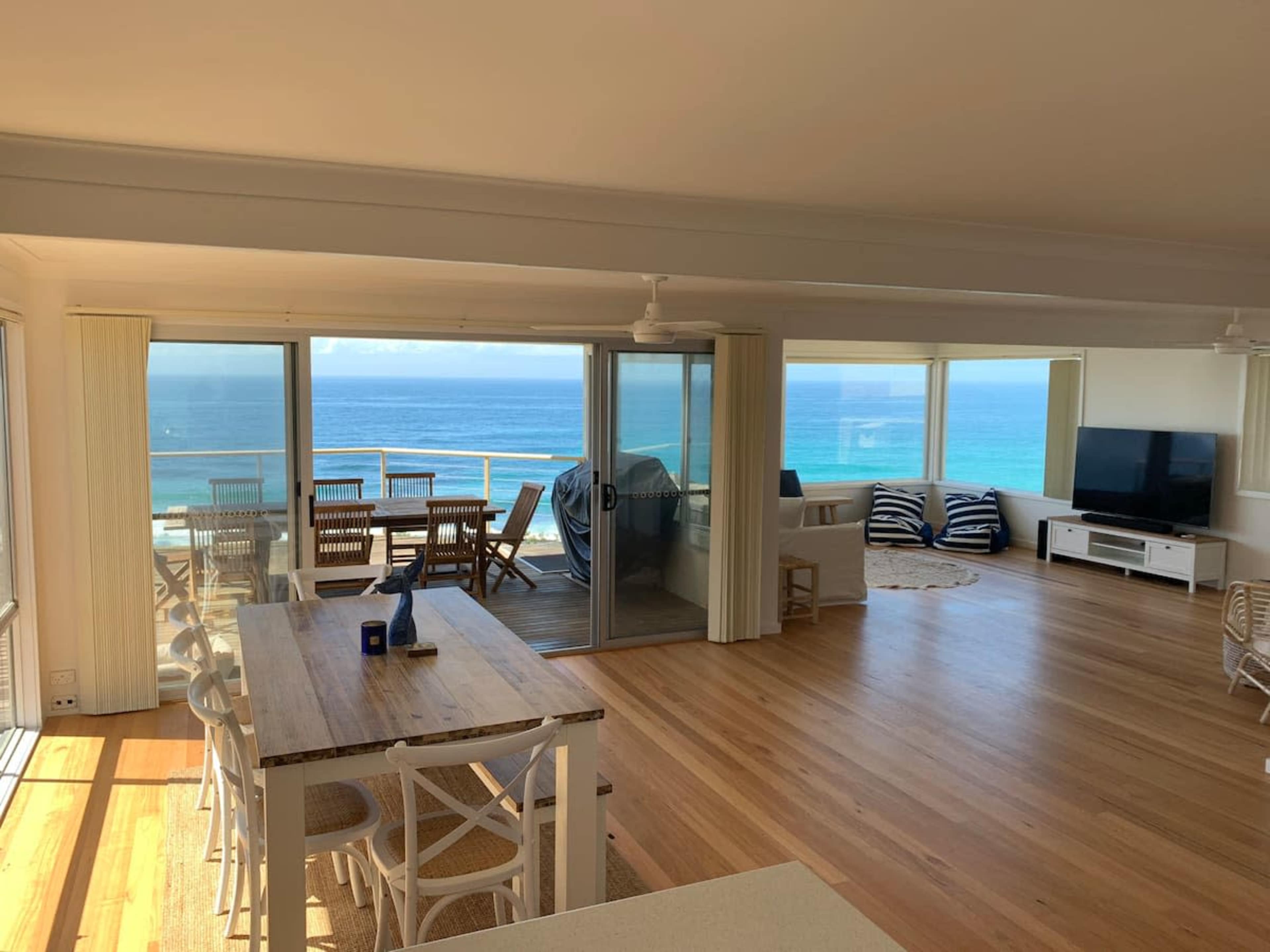 A modern living space with large windows overlooking an ocean view, featuring a dining area with a wooden table and chairs, a comfortable seating area, and light-colored wooden flooring.