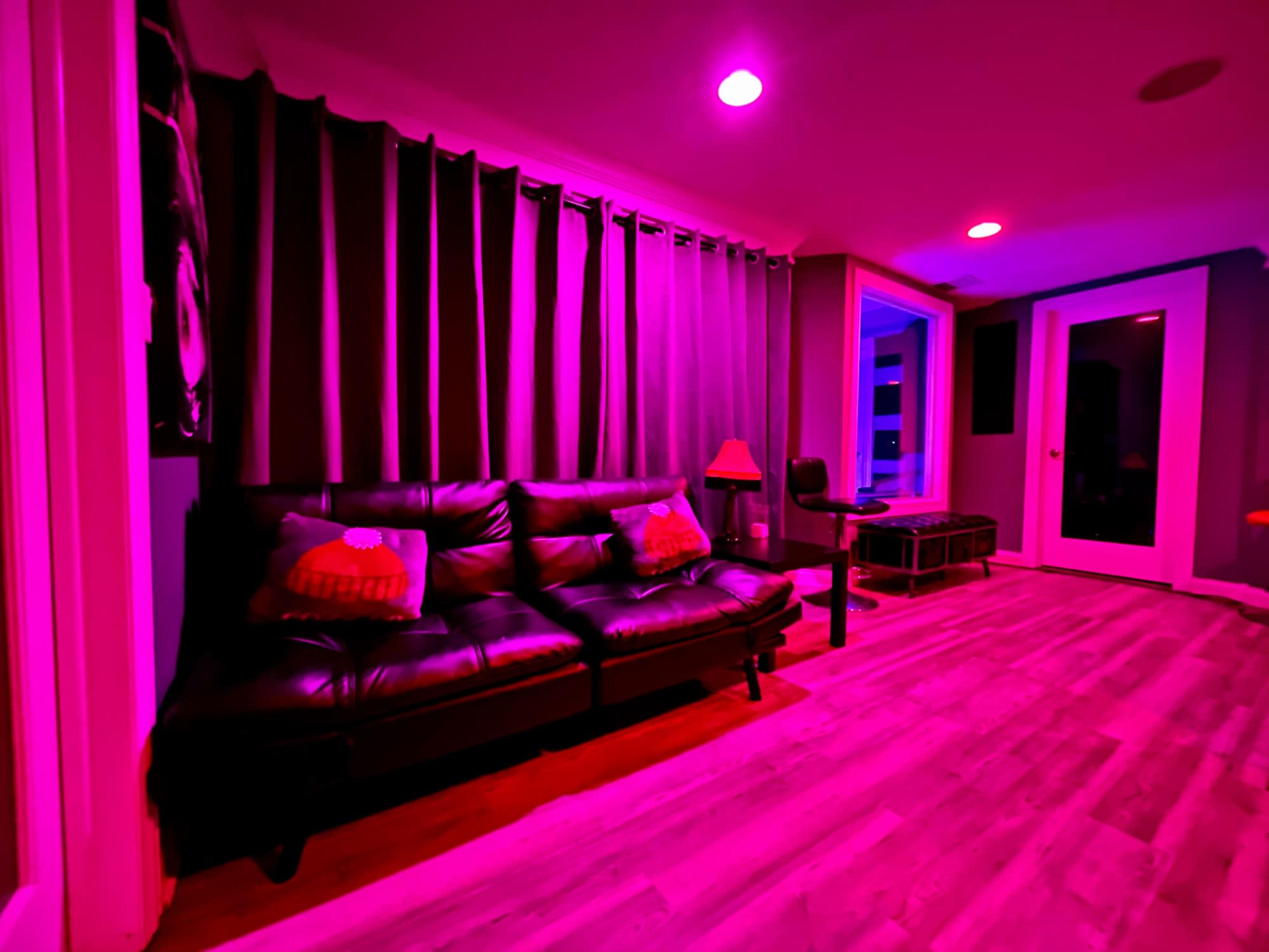 A modern living room features a black couch with pillows, a small lamp, and purple lighting, alongside curtains and large windows.