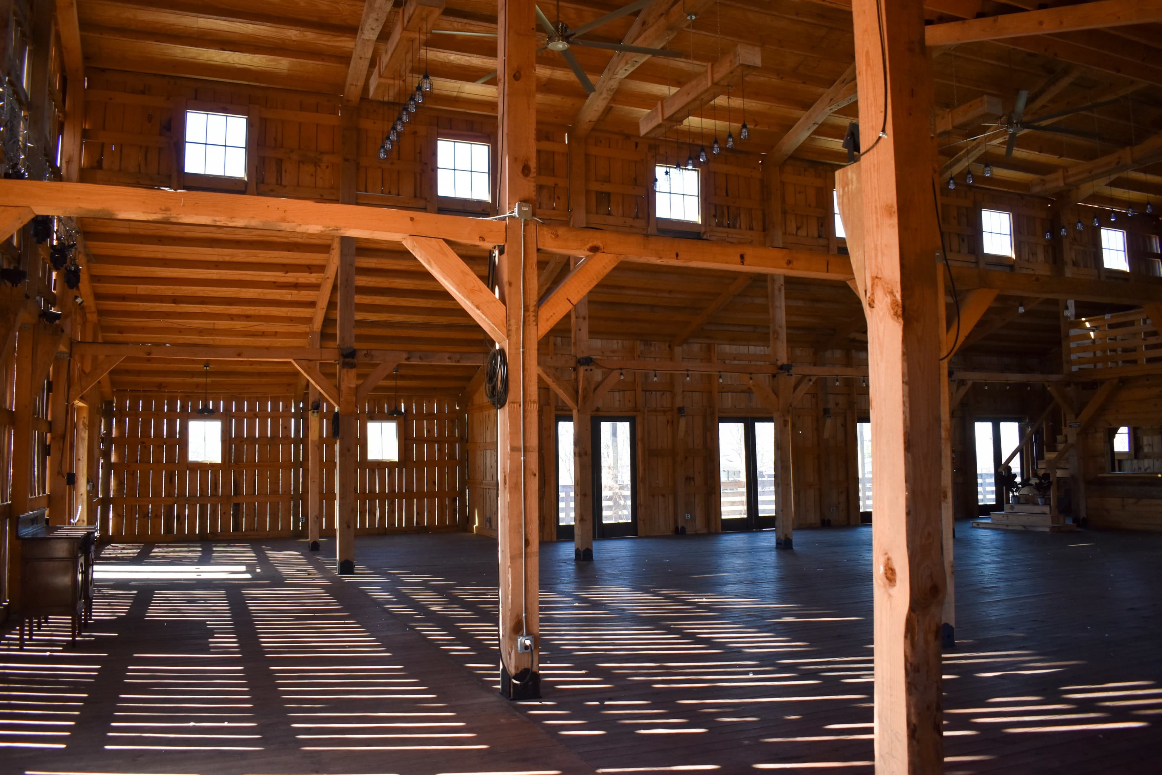 The interior of a wooden barn with large beams, tall ceilings, and sunlight casting shadows on the floor through the windows.