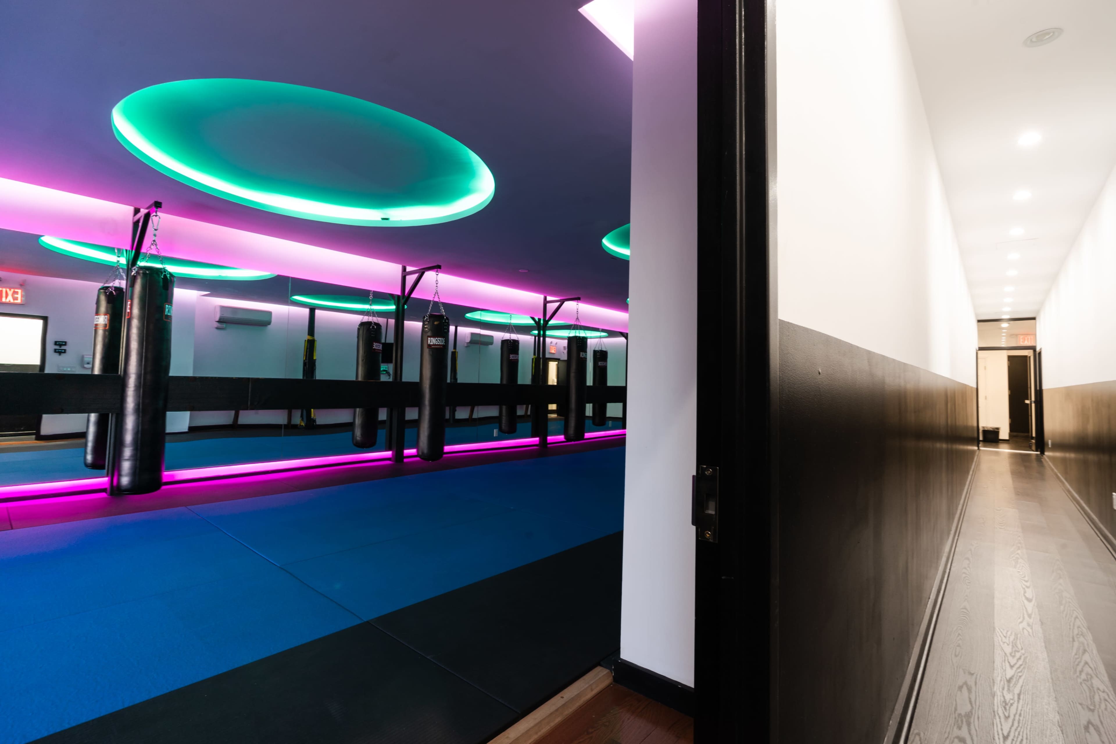 A modern gym interior featuring a row of punching bags illuminated by colorful LED lights, with a hallway visible in the background.