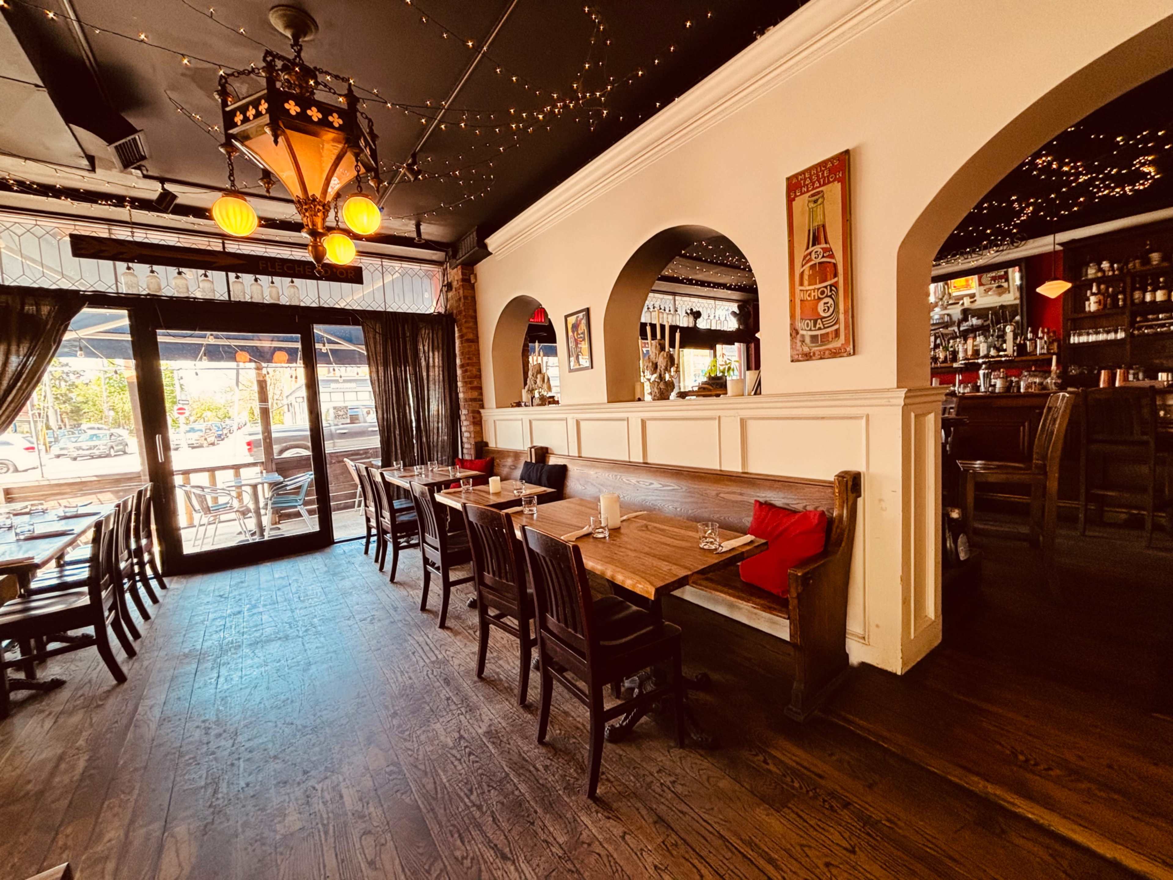 The image shows an interior view of a restaurant featuring wooden tables, chairs, and decorative elements, with a bar visible in the background.