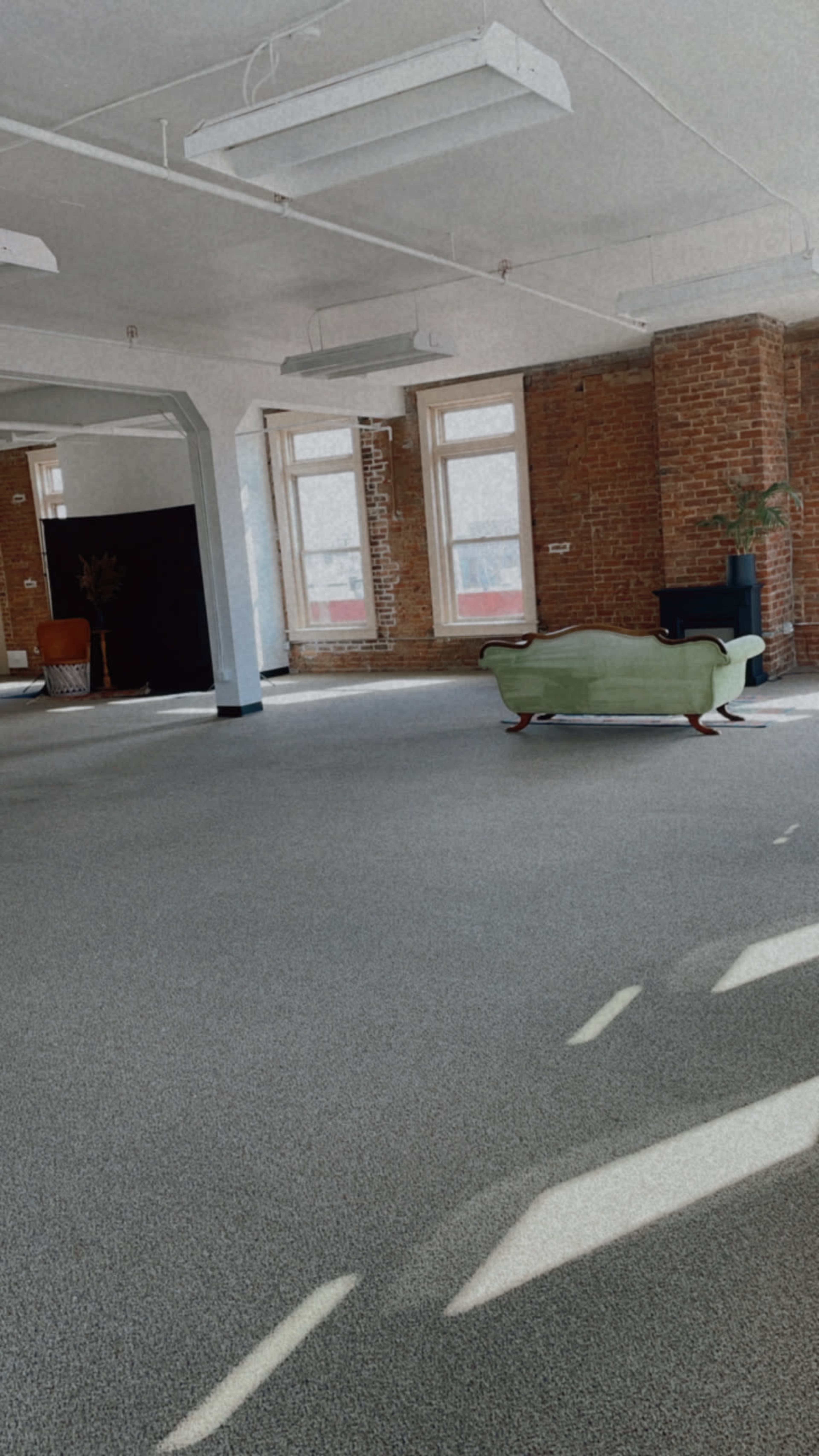 The image shows a spacious, empty room with brick walls, large windows, and a green couch positioned in the center.