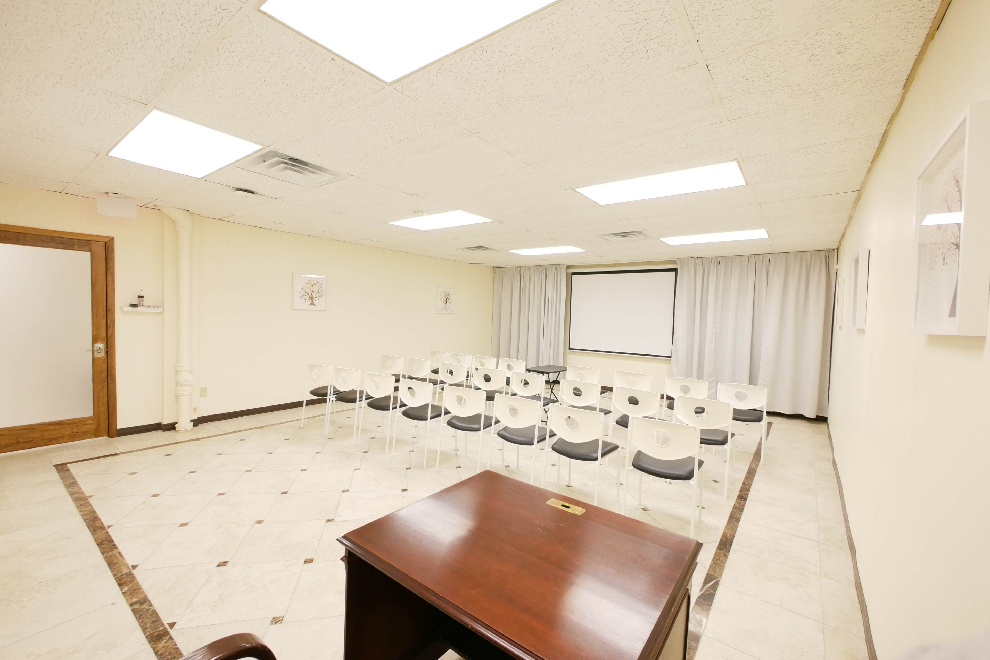 The image shows a meeting room with rows of white chairs facing a screen, a wooden desk in the foreground, and a light-colored tiled floor.
