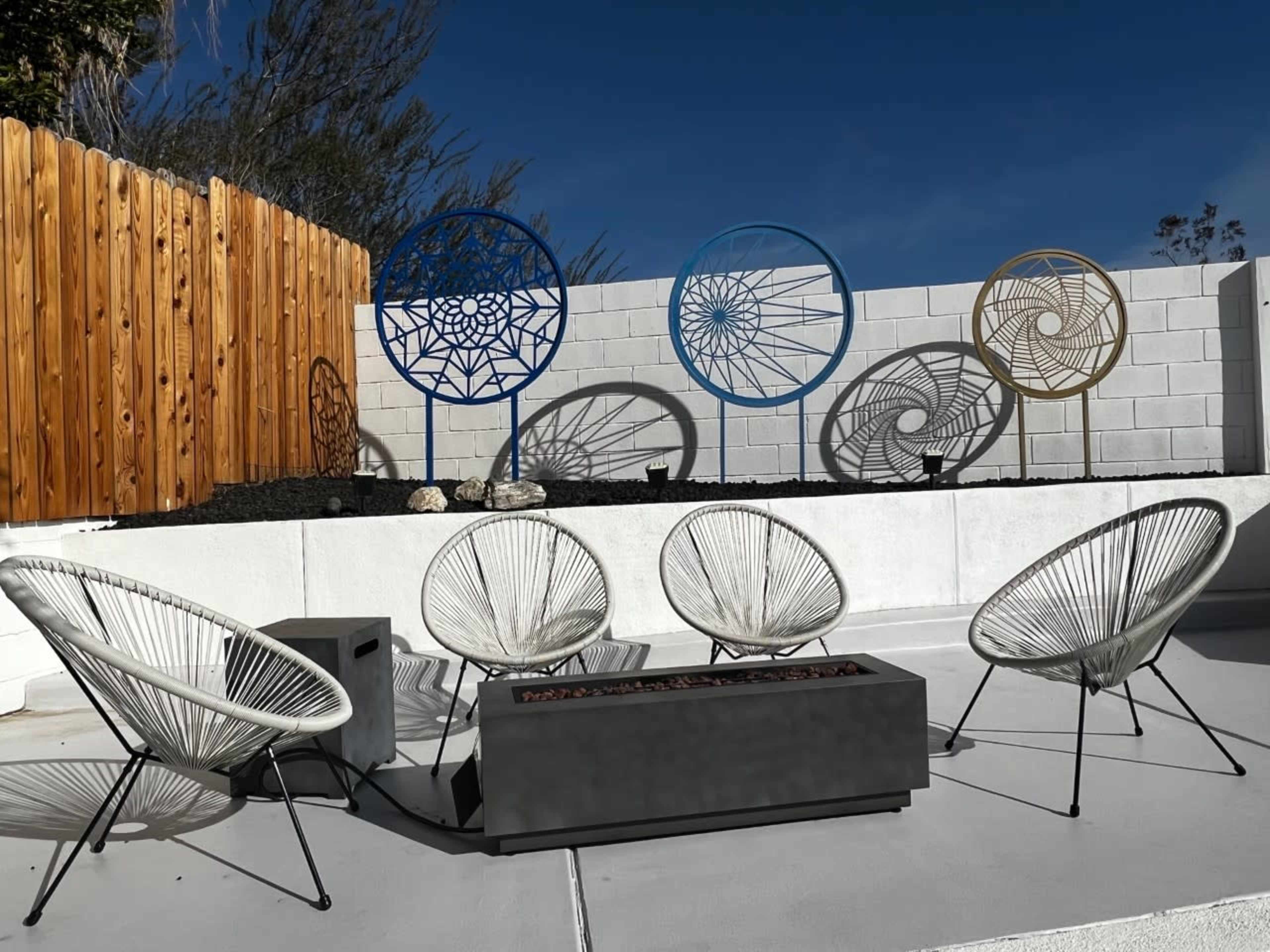 The image shows a patio area with five Acapulco chairs arranged around a rectangular fire pit, set against a white wall featuring decorative geometric wall art.