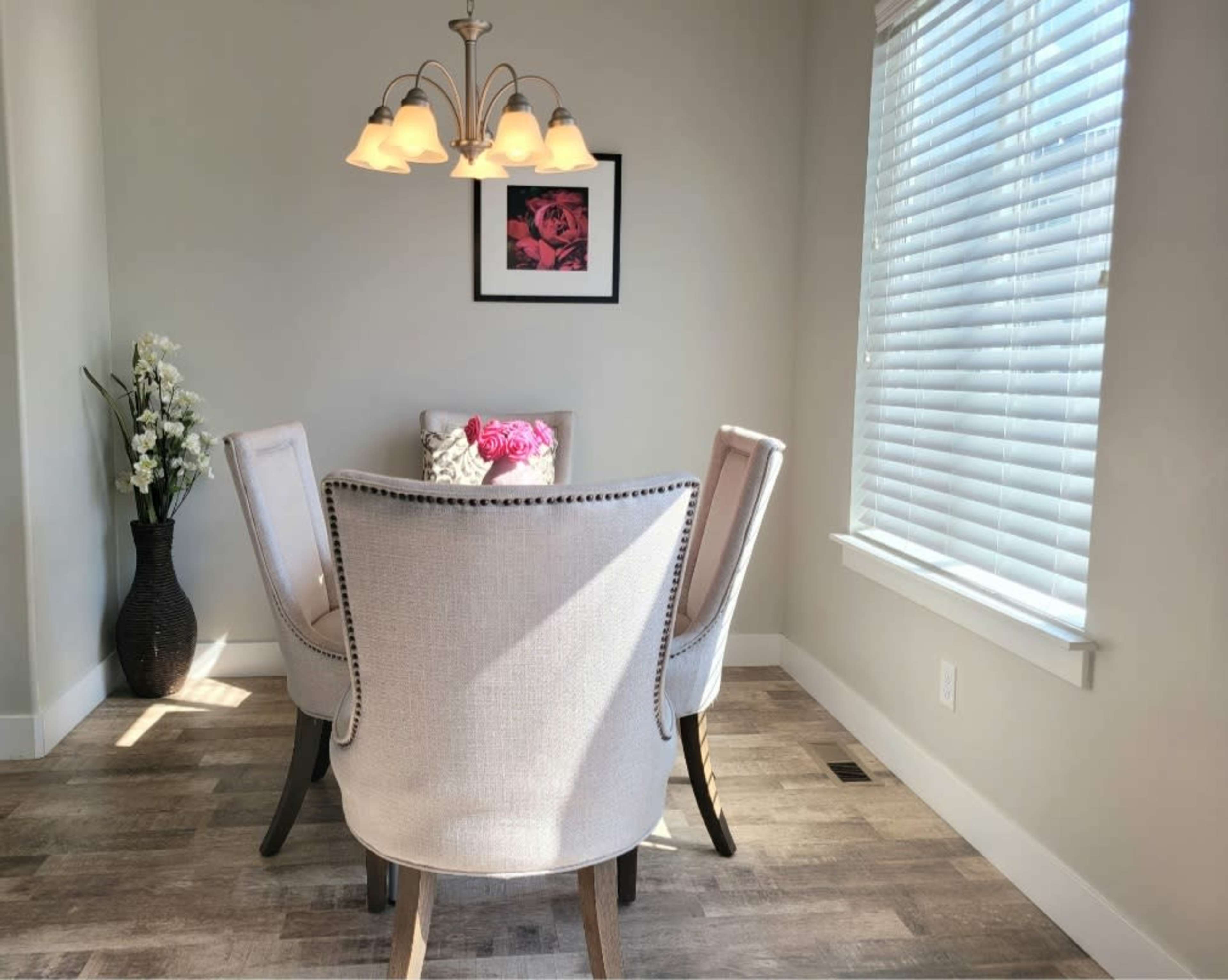 A dining area features a round table with four upholstered chairs, a floral centerpiece, and a framed picture on the wall.