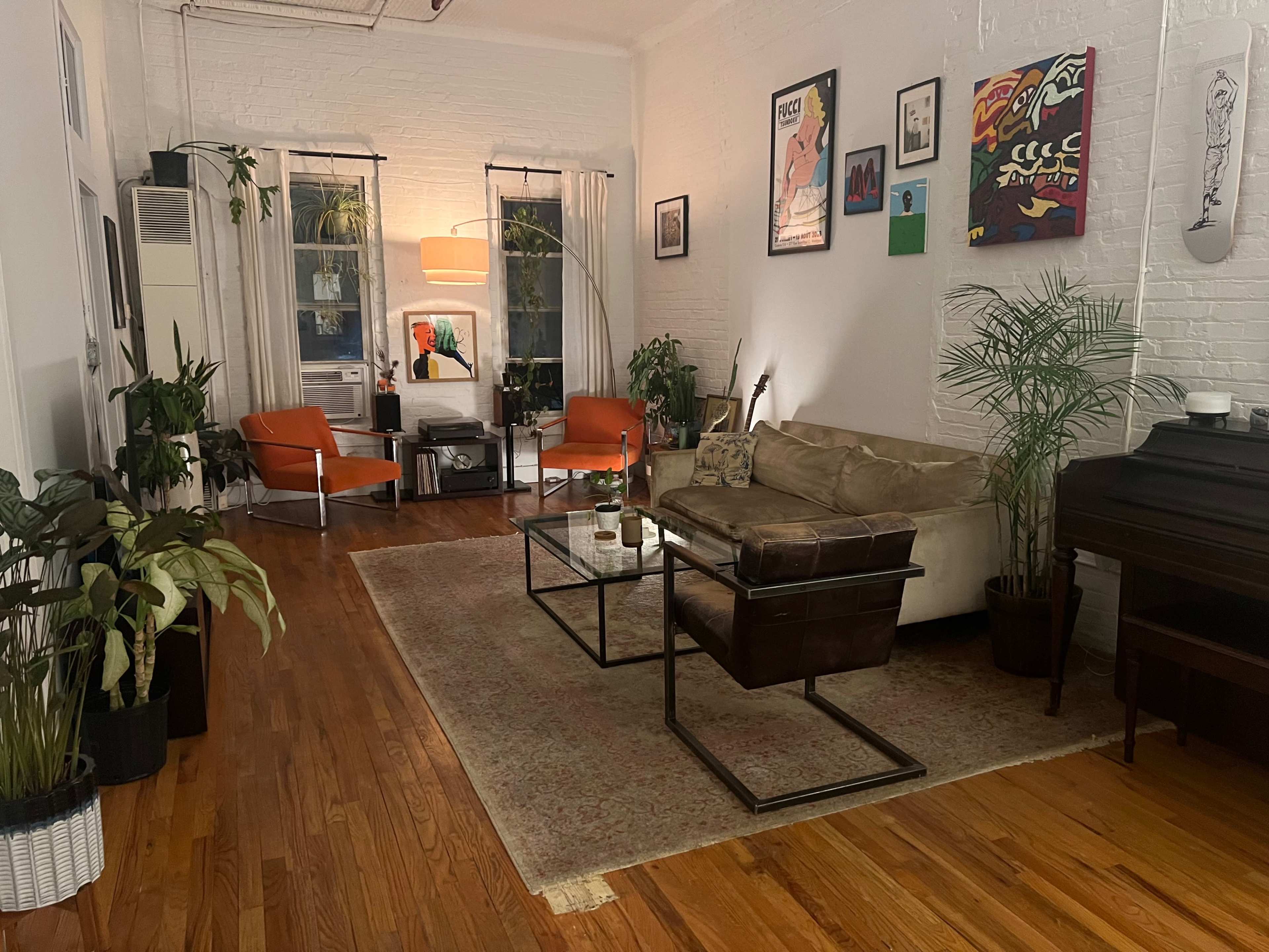 The image shows a cozy living room with a sofa, two orange chairs, a coffee table, plants, and framed artwork on the walls.
