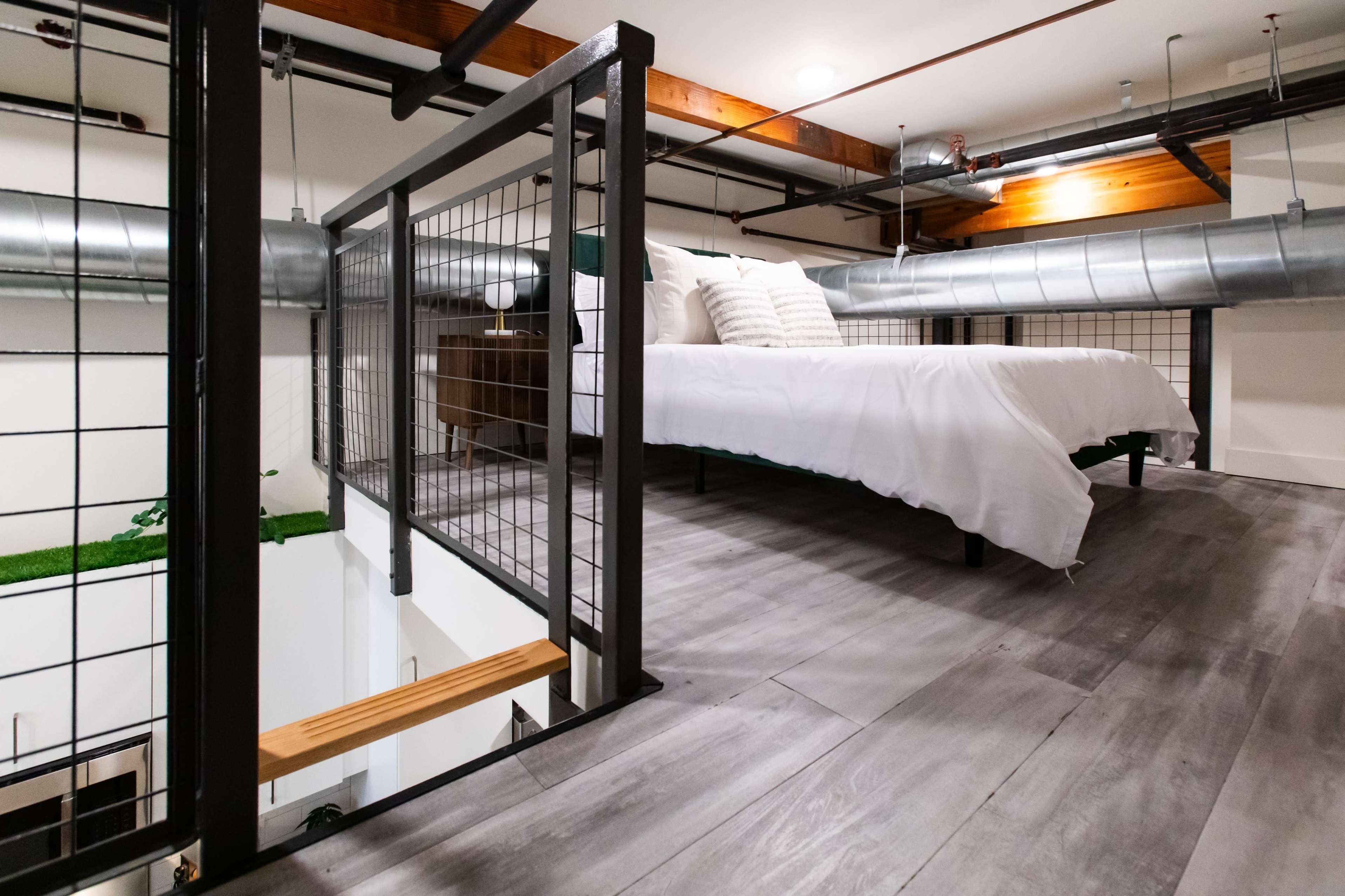 A lofted bedroom with a bed on a wooden platform surrounded by metal railings and industrial-style ductwork overhead.