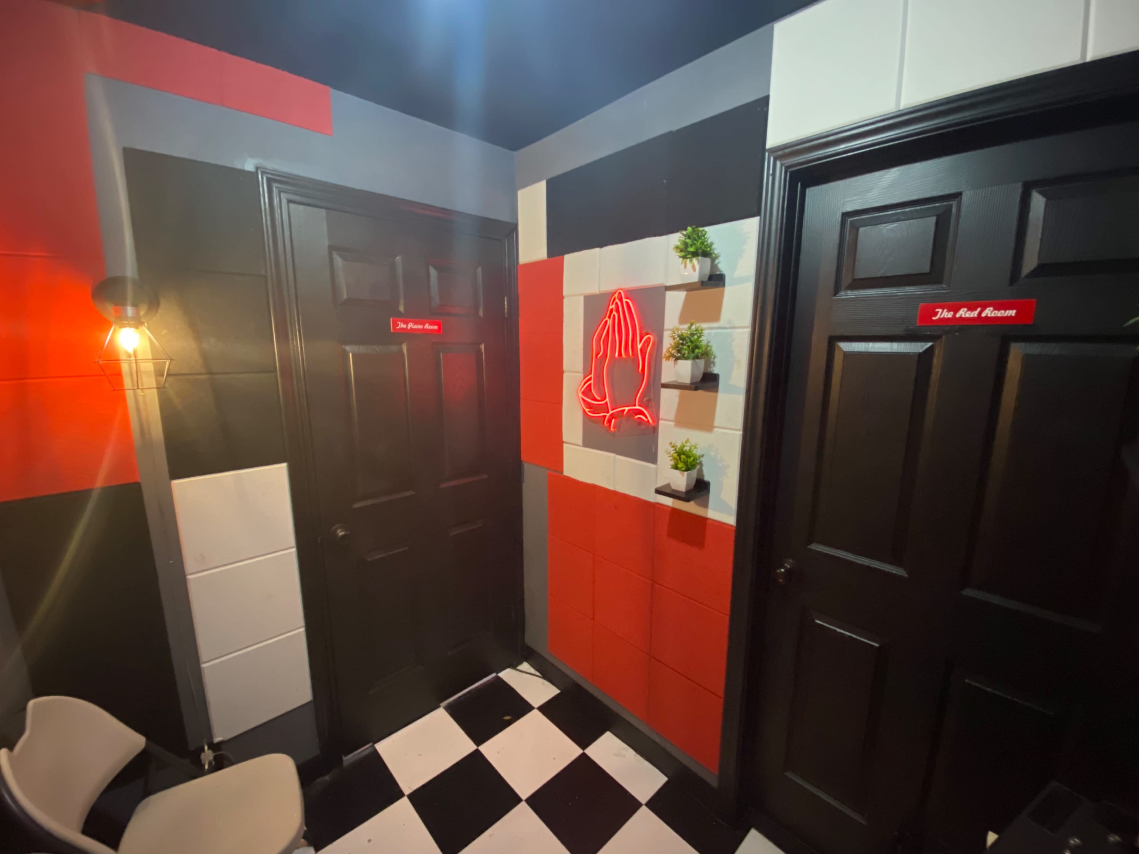 A corner of a room with two black doors, colorful walls featuring red and black geometric patterns, and a neon sign of praying hands alongside small plants.