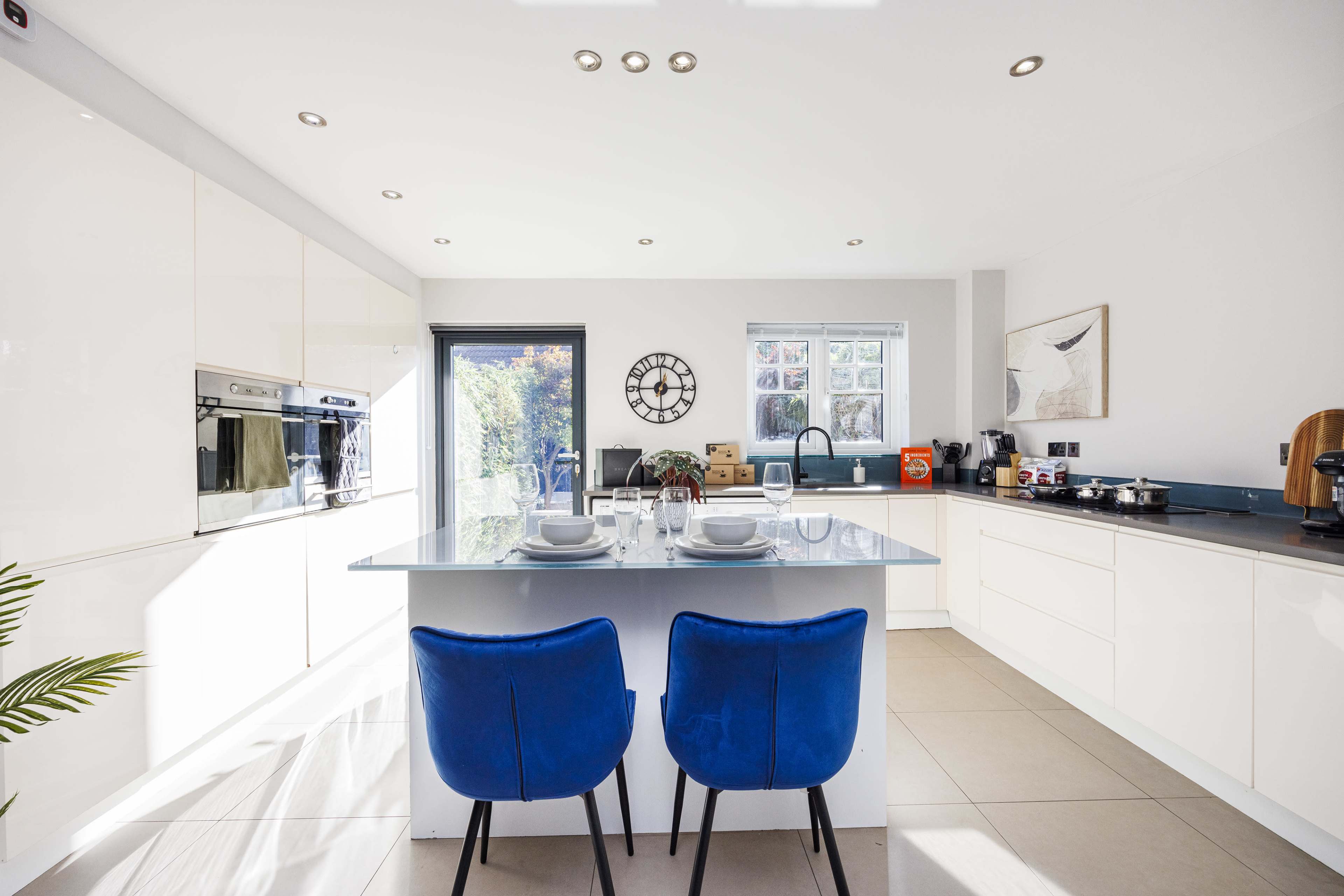 A modern kitchen features a central island with two blue chairs, white cabinets, and large windows letting in natural light.