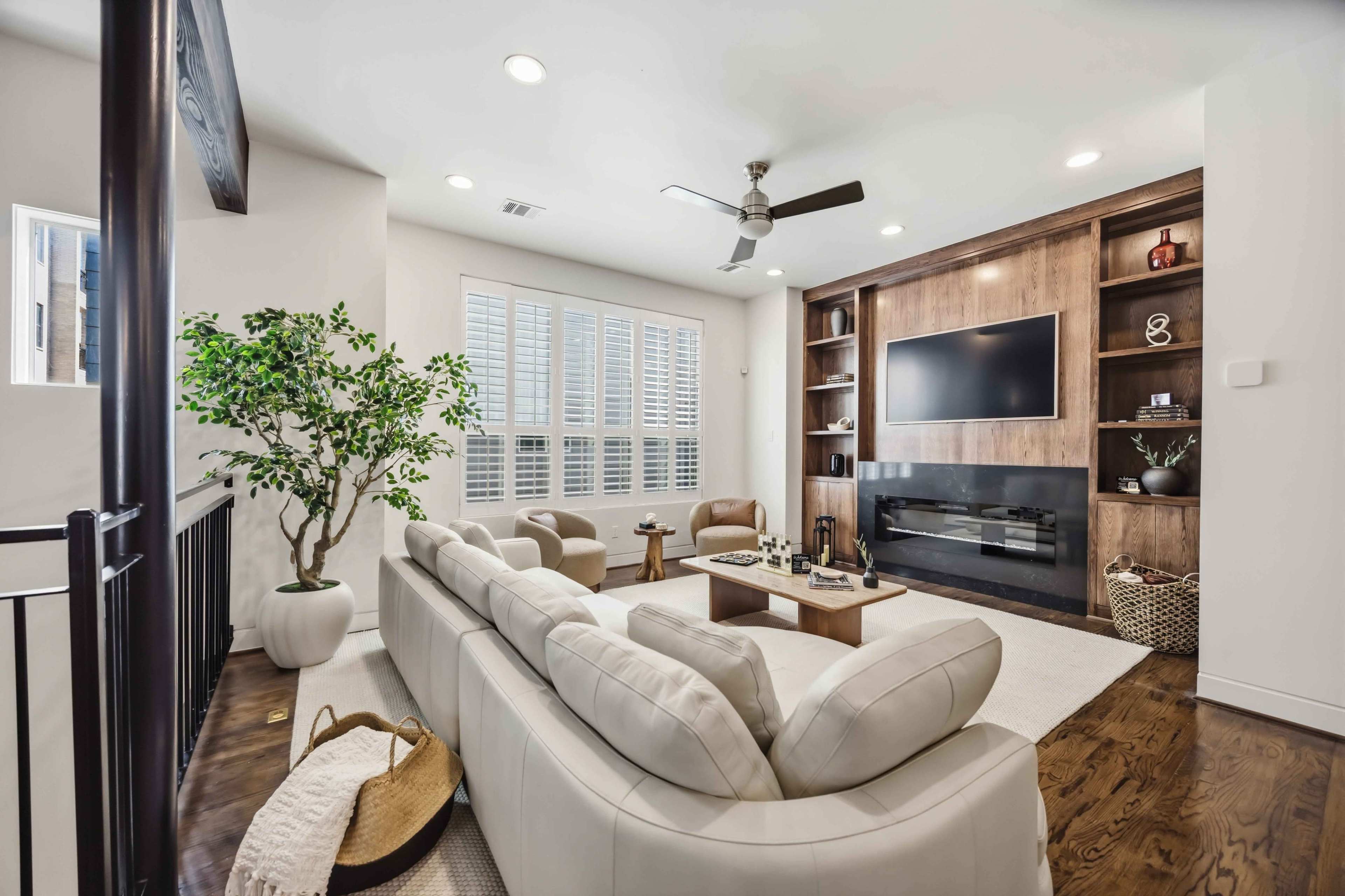 The image shows a modern living room featuring a light-colored leather sectional sofa, a wooden entertainment unit, and a large window with white shutters.