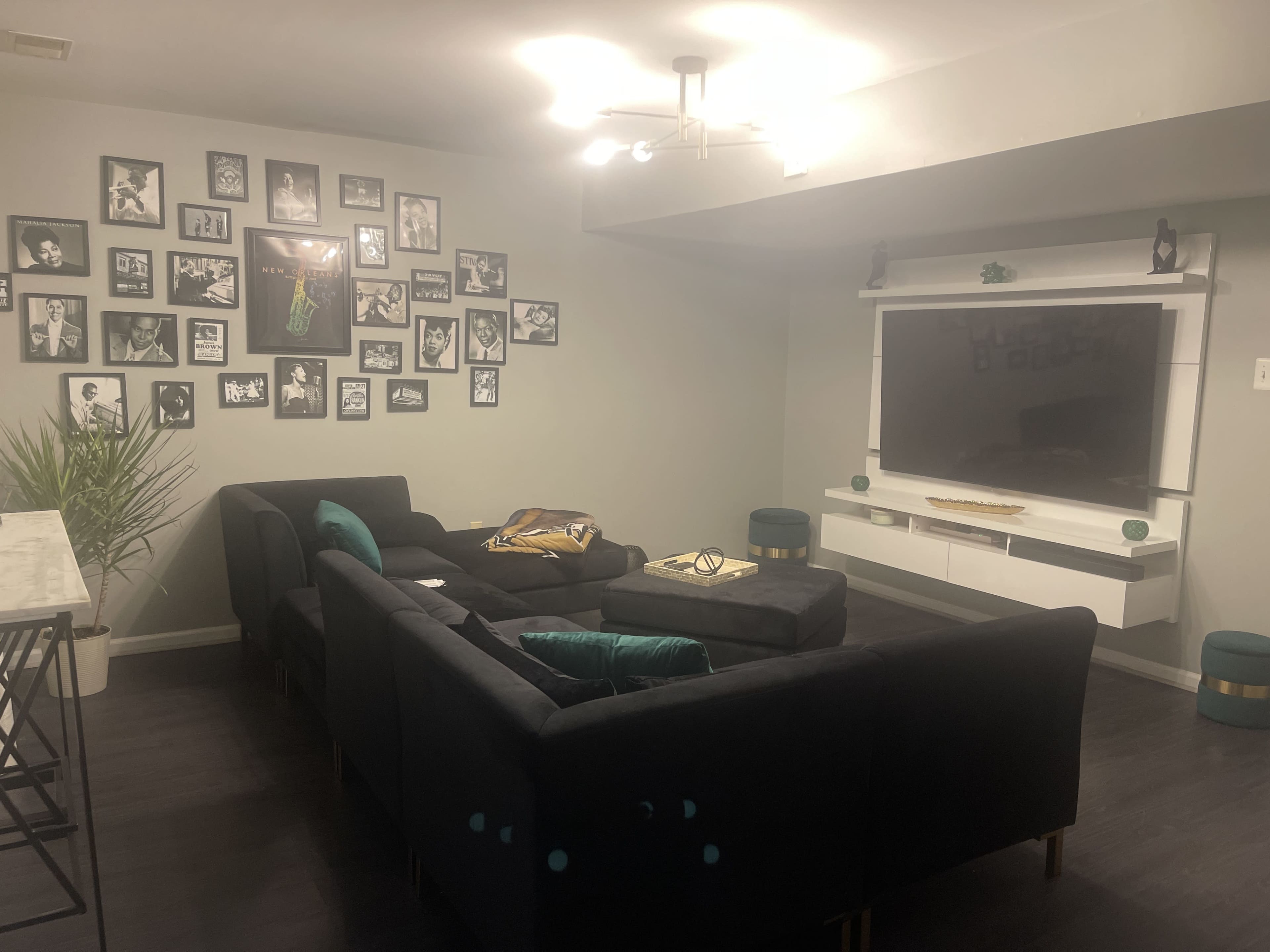 The image shows a living room with a dark sectional couch, a wall adorned with framed black-and-white photographs, a large television mounted on the wall, and a few decorative green accents.