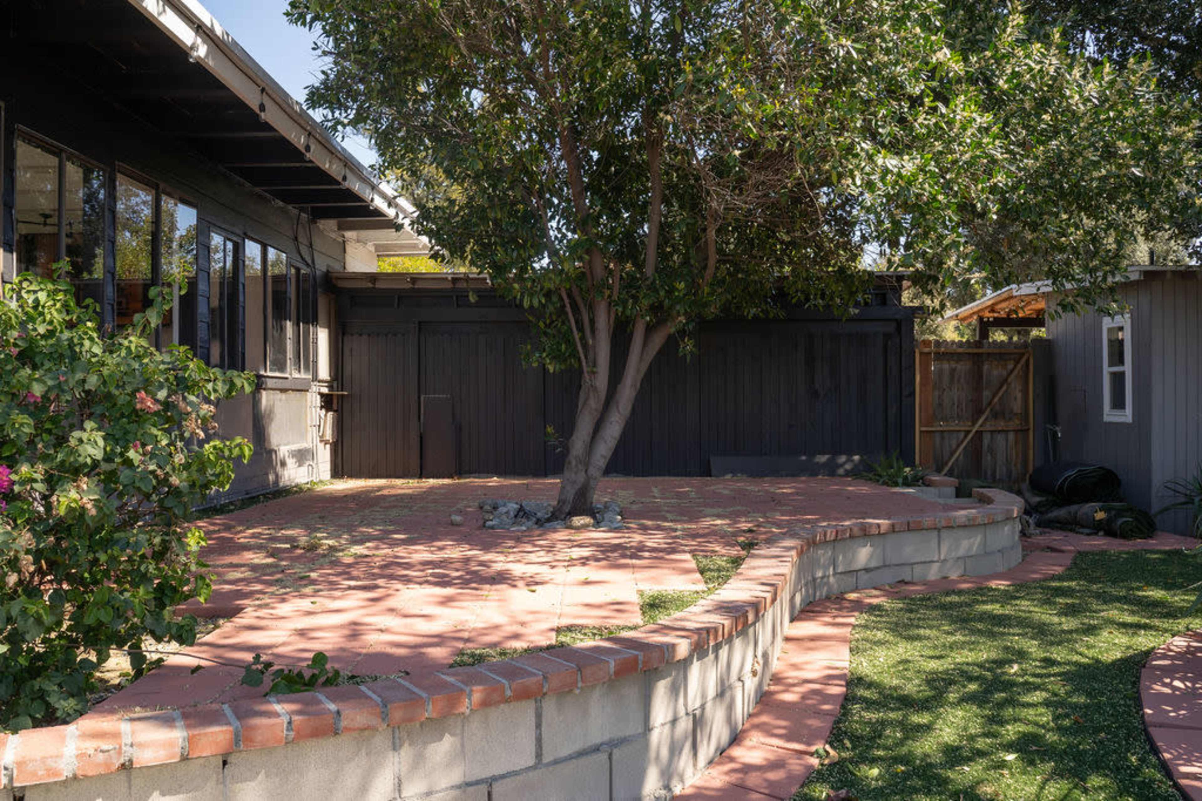 The image shows a landscaped outdoor area featuring a brick-bordered pathway, a tree, and a wooden fence in the background.