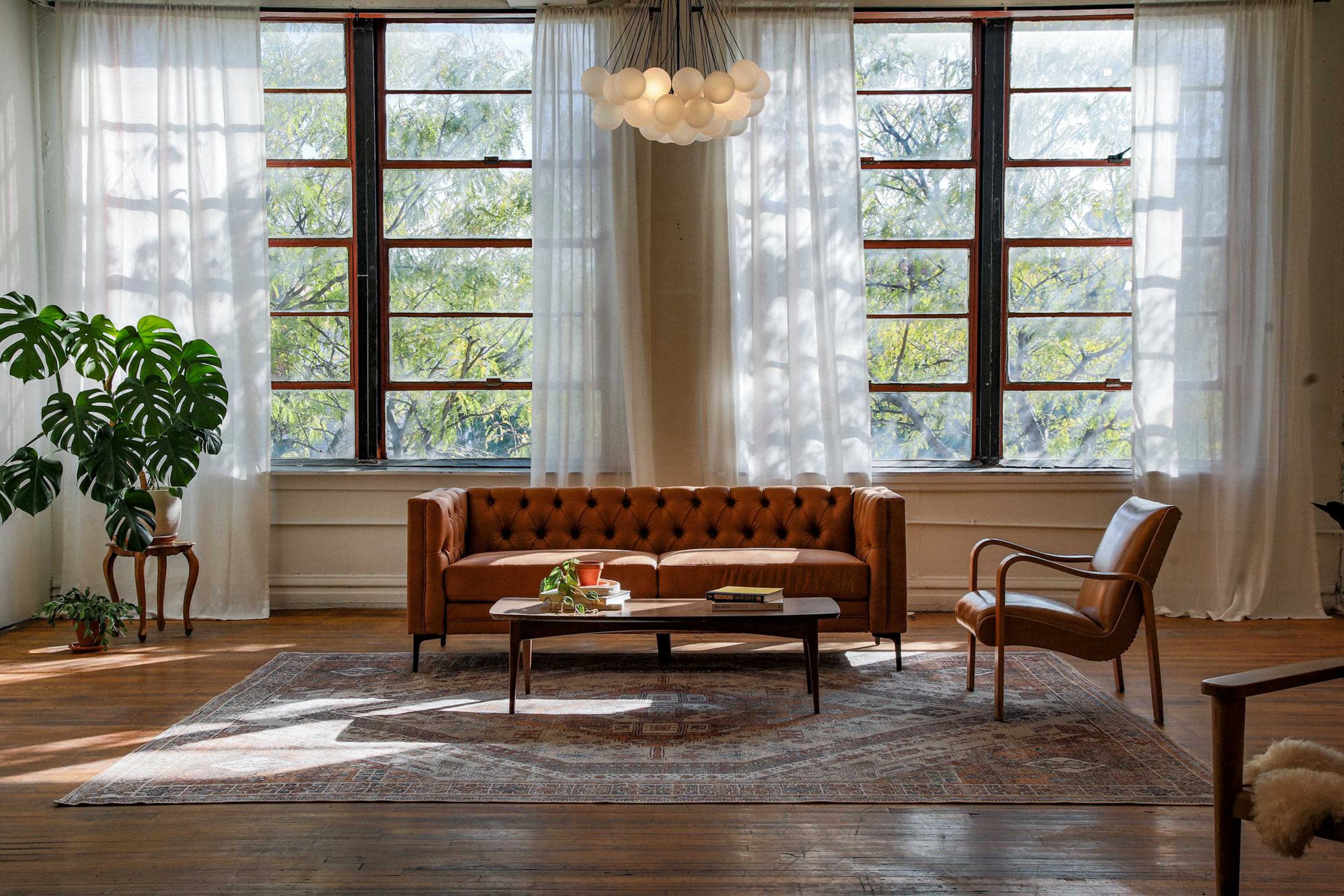 A sunlit living room featuring a brown leather couch, a wooden coffee table, and a single armchair, with large windows allowing greenery to be visible outside.