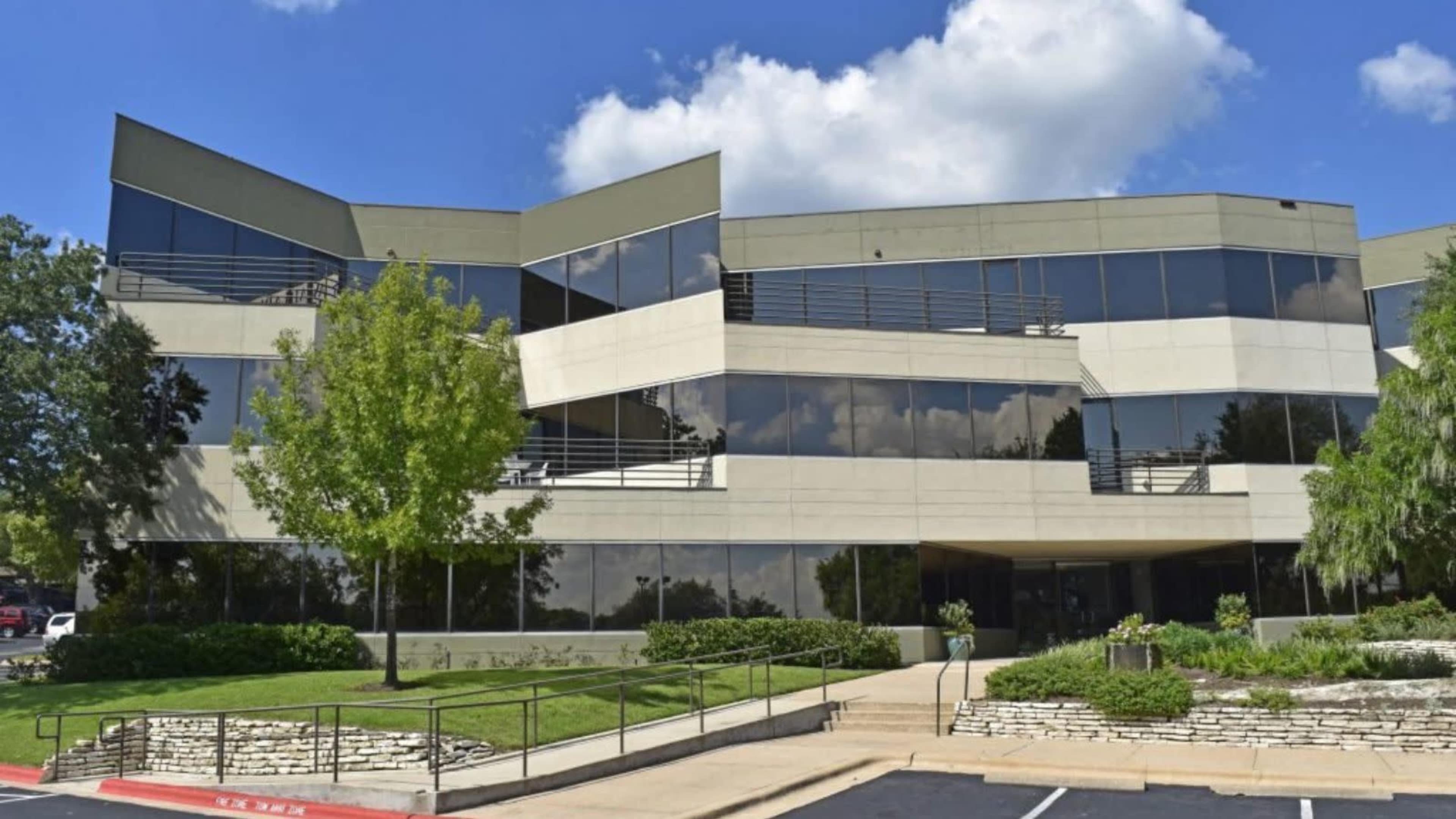 The image shows a modern, multi-level office building with large windows and landscaped surroundings.