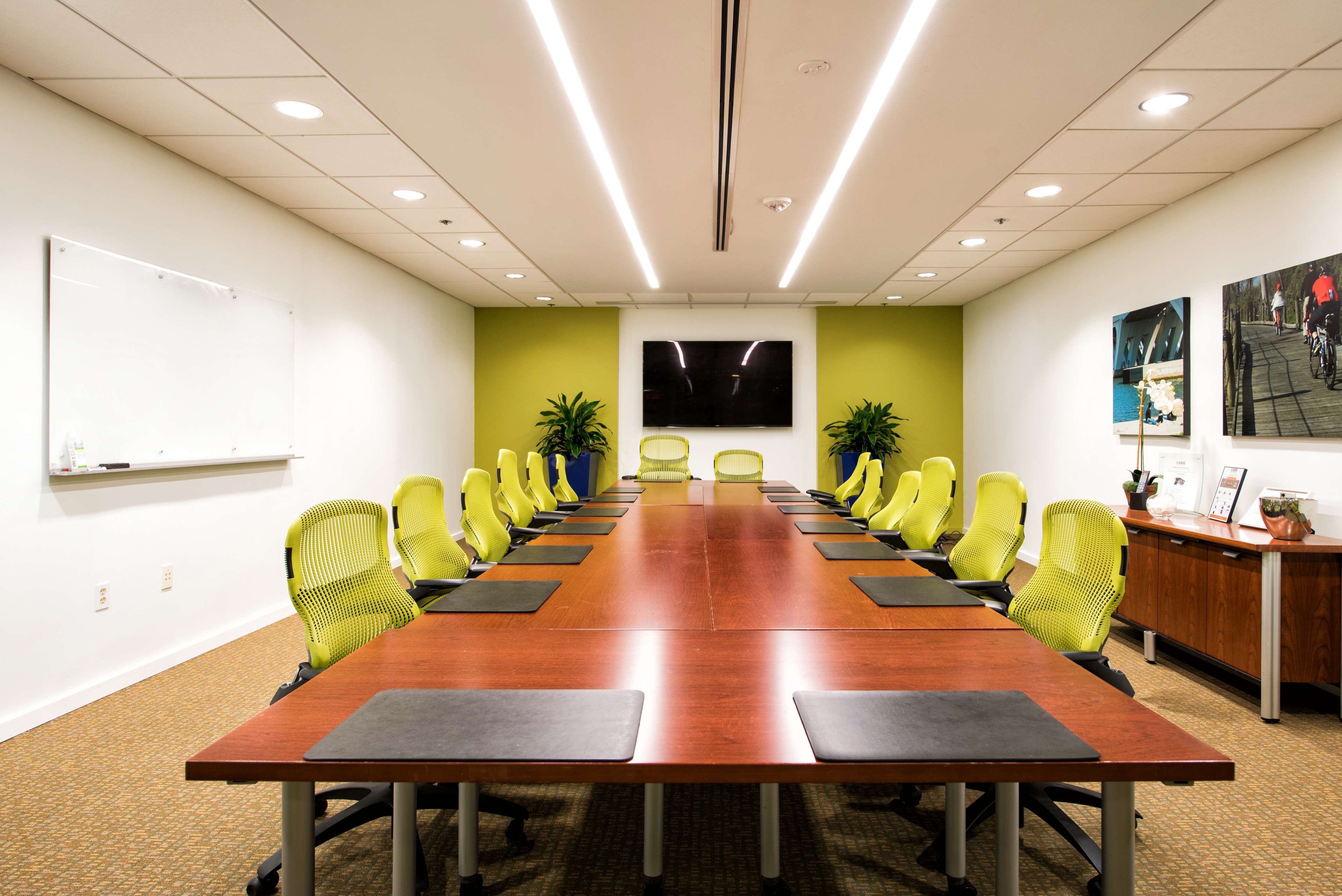 A long wooden conference table is set in a modern meeting room with green accent walls and ergonomic yellow chairs.