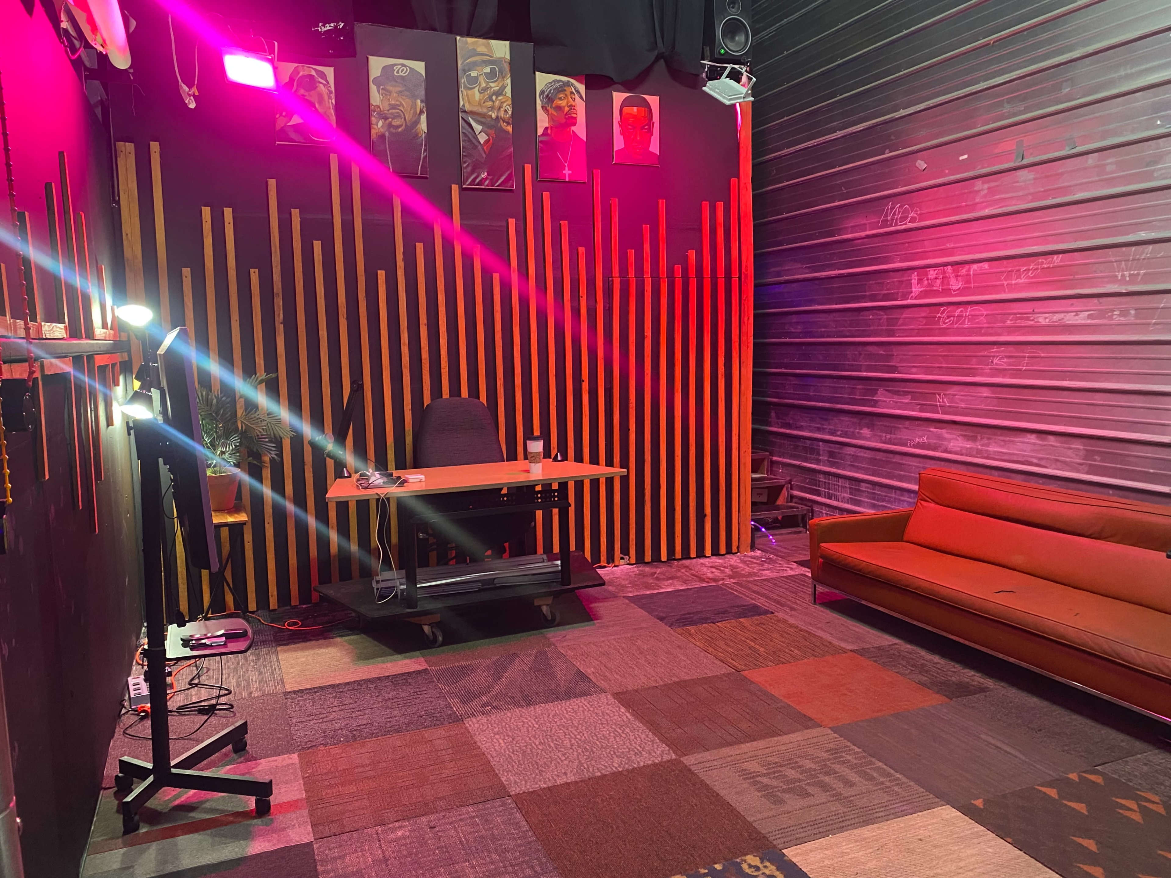 The image shows a modern, sparse office space with a wooden desk, a chair, and a retro orange couch, all set against a wall adorned with vertical wooden panels and framed artworks.