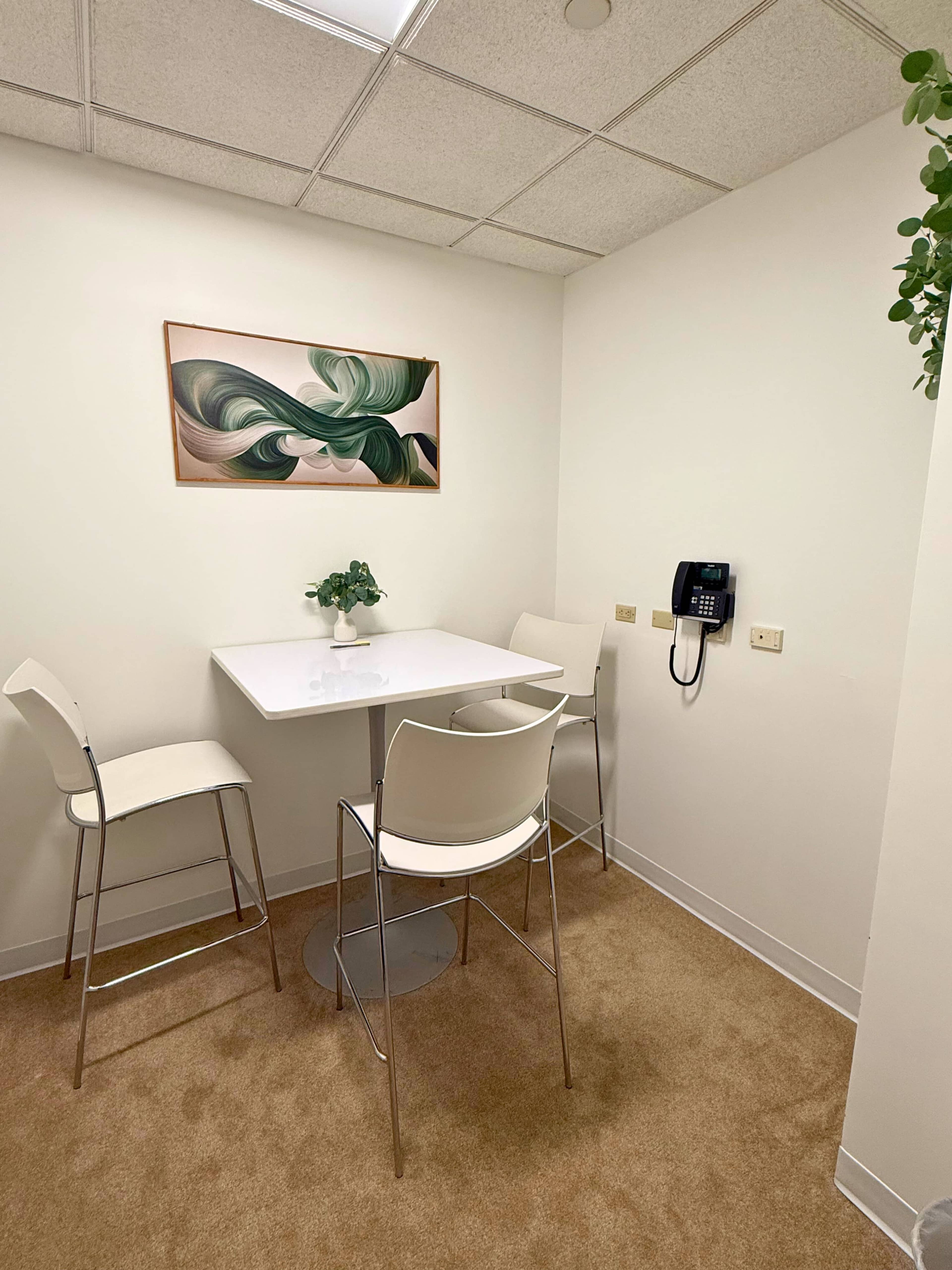 The image shows a small meeting space with a white table, four high chairs, a potted plant on the table, a wall-mounted telephone, and abstract artwork on the wall.