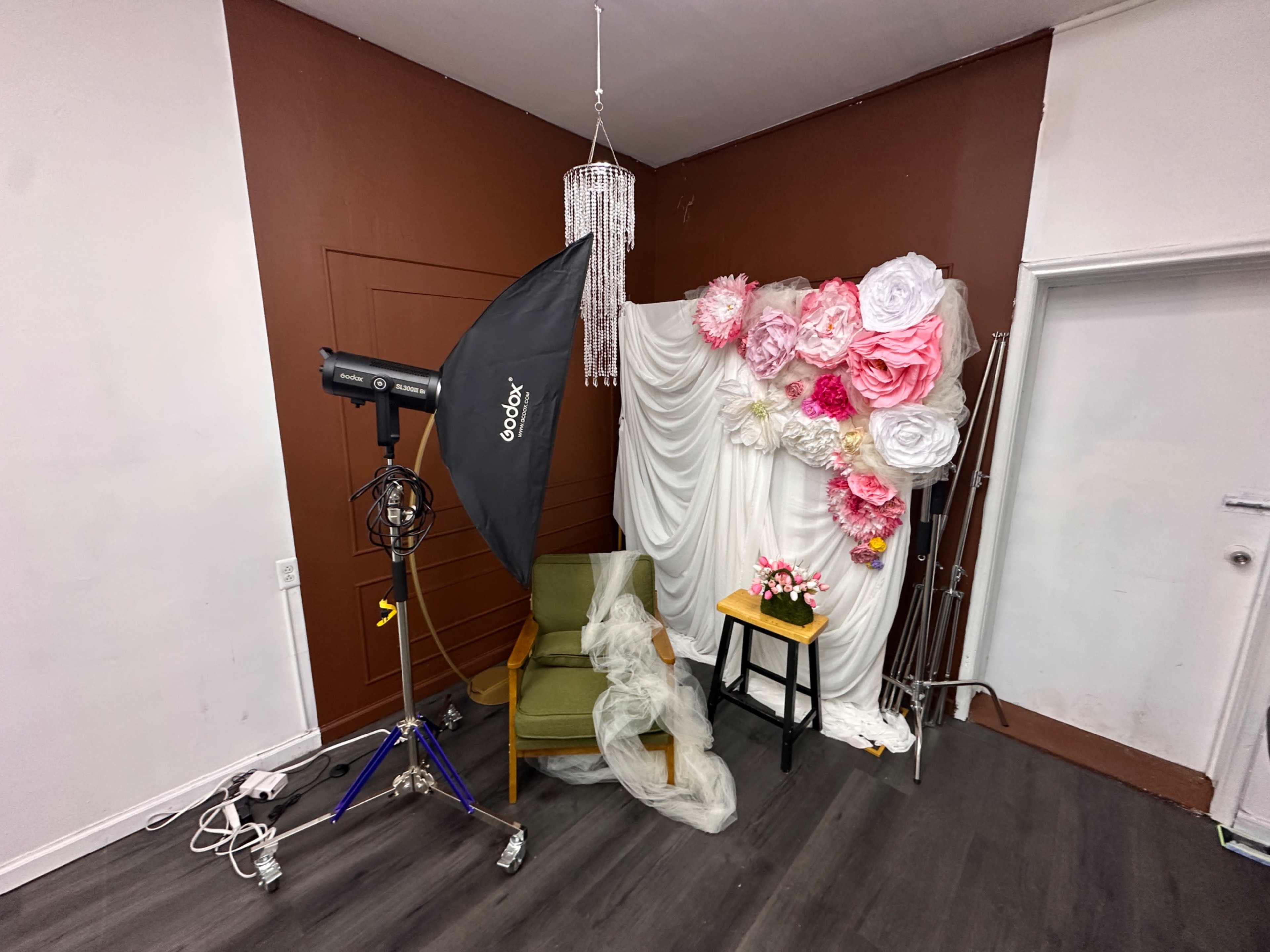 The scene shows a photography setup with a green chair, a small black table with flowers, a softbox light, and decorative fabric and paper flowers against a brown wall.