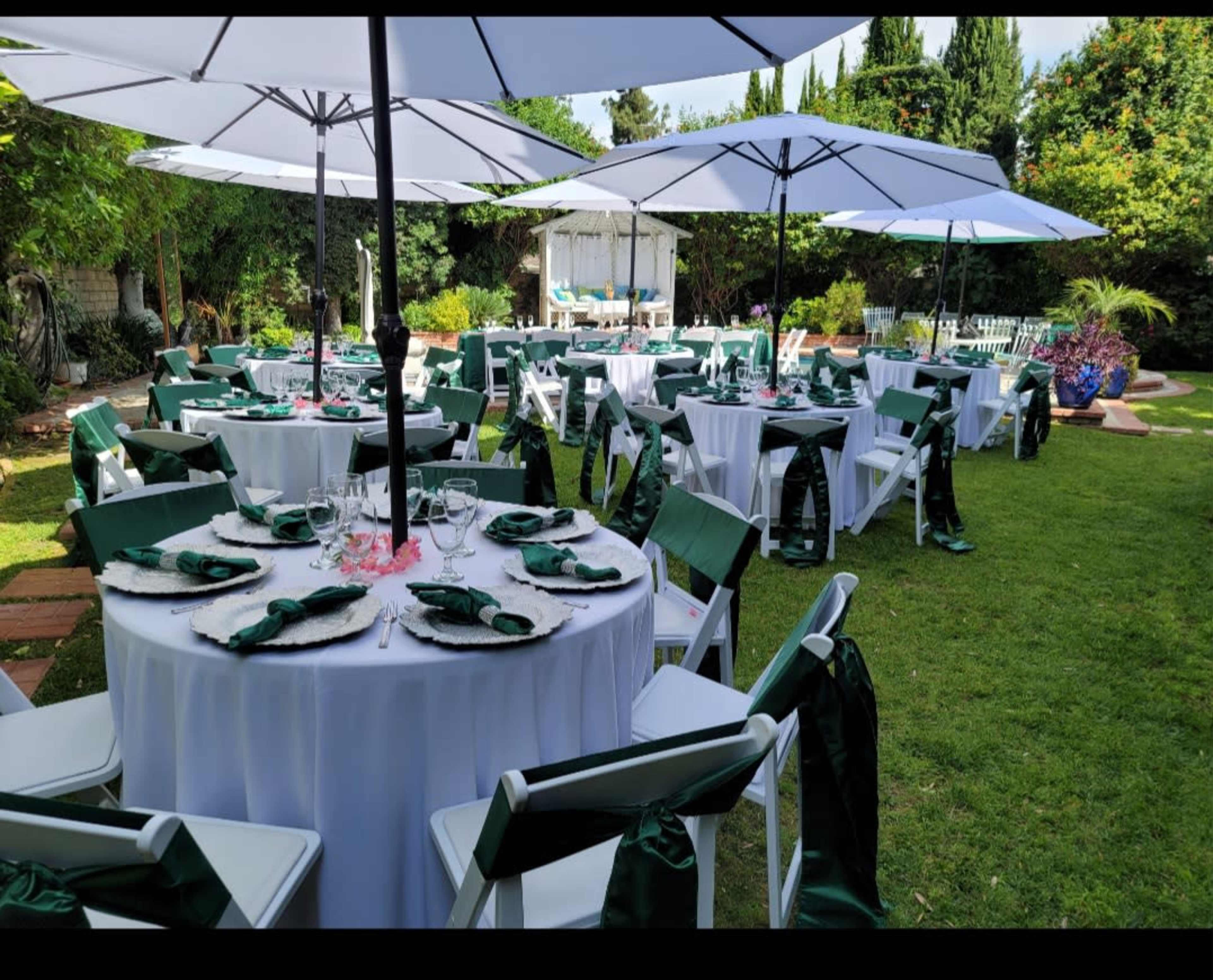 The scene features a well-decorated outdoor garden setting with tables covered in white linens and green accents, surrounded by lush greenery and large umbrellas.